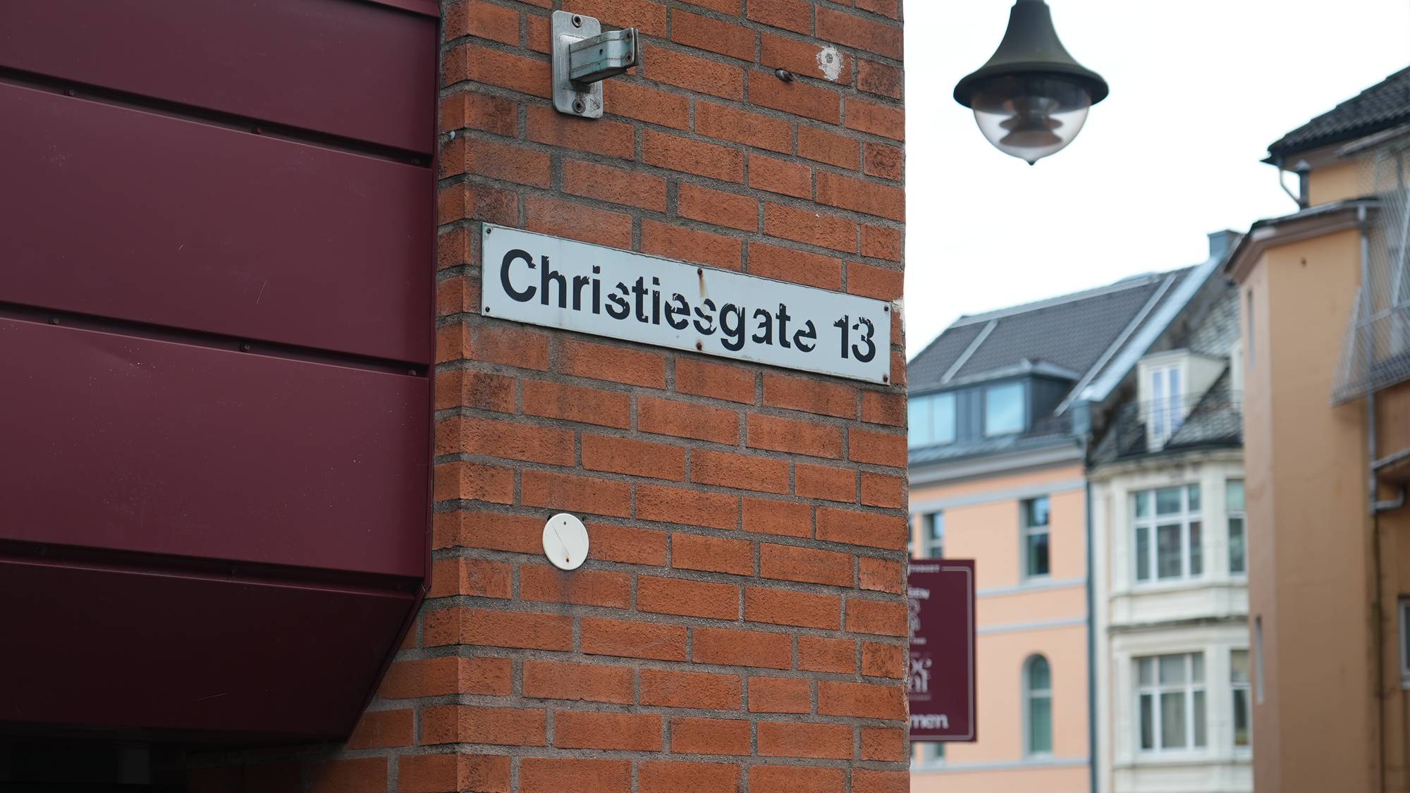Gatelykt over en mursteinsvegg. Gatenavnet "Christiesgate 13" er synlig på et hvitt skilt. I bakgrunnen ses fargerike bygårder. Det er en person med grått hår som ses fra siden, i forgrunnen til venstre. (Bildebeskrivelsen er laget av en KI-tjeneste)