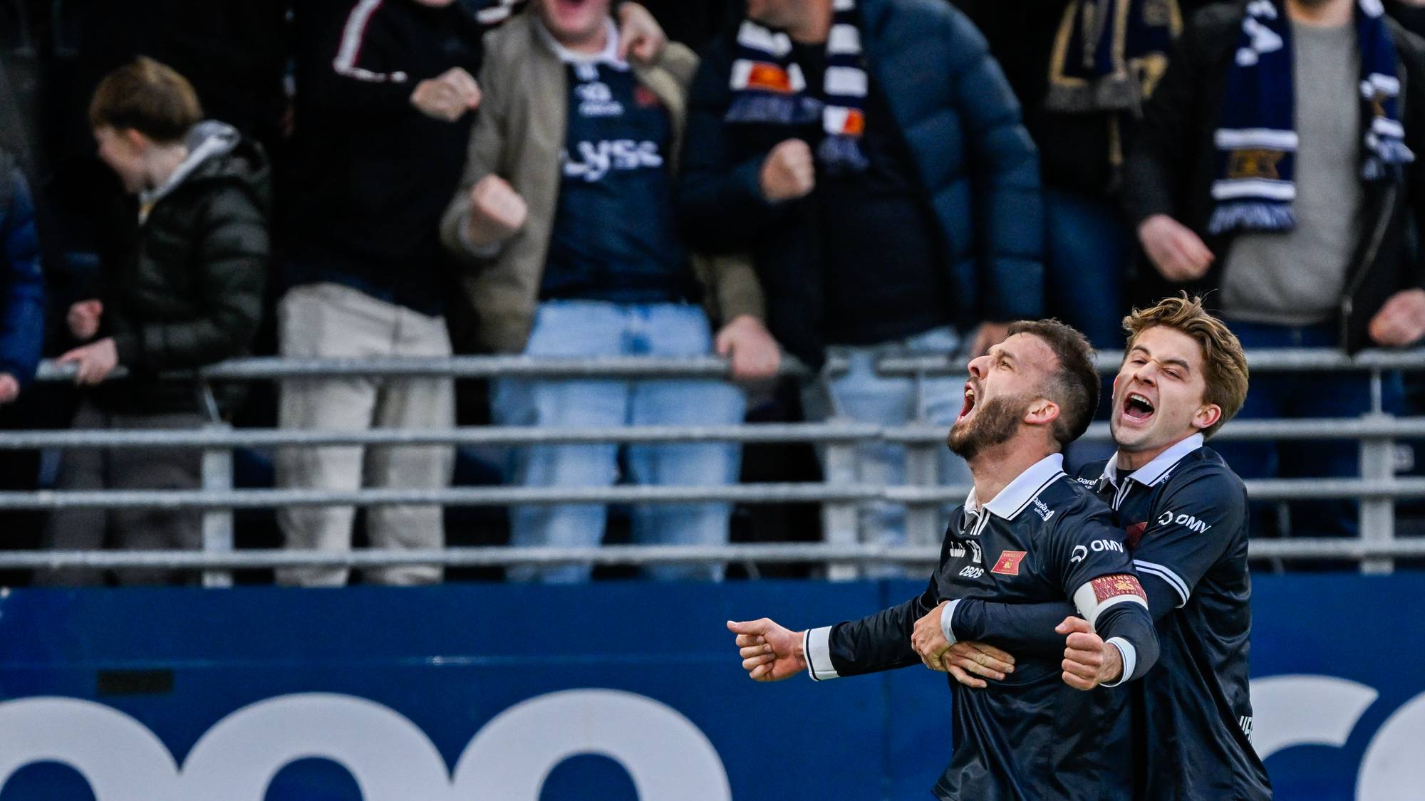 To fotballspillere feirer et mål under en kamp. Begge er kledd i mørke draktsett. I bakgrunnen sitter publikum på tribunen. (Bildebeskrivelsen er laget av en KI-tjeneste)