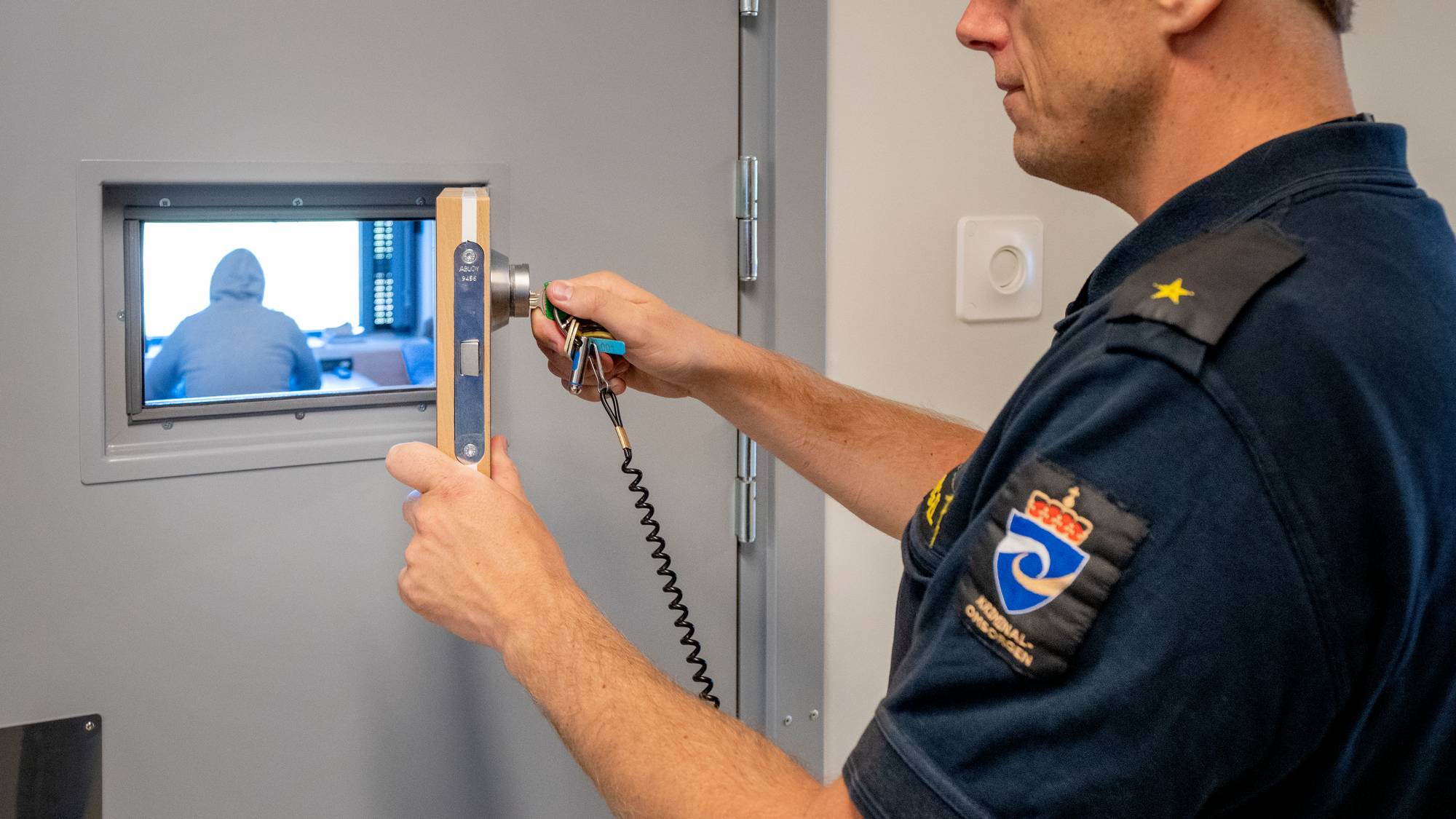 Fengselsbetjent låser celledør med innsatt synlig gjennom vinduet. 