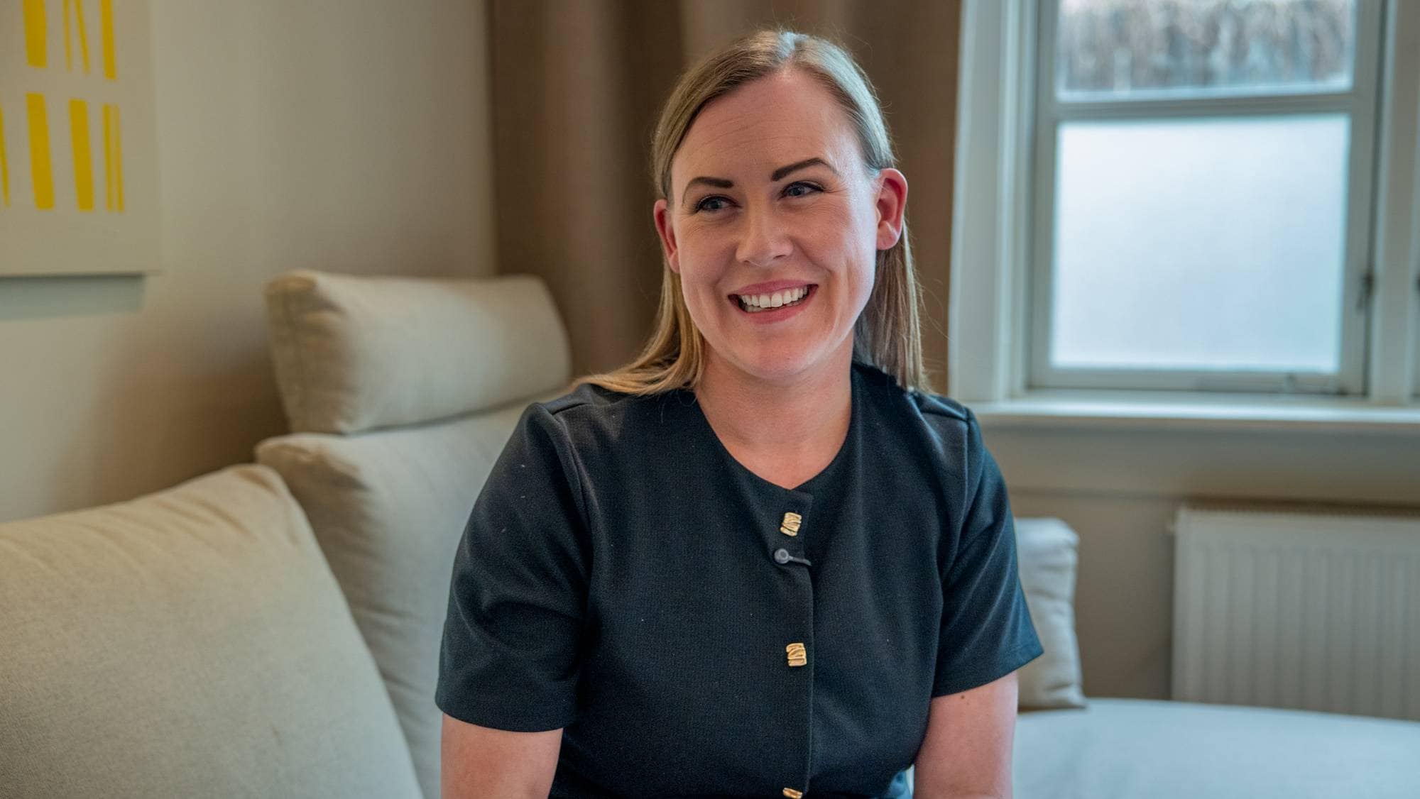 En kvinne med blondt hår sitter i en lys sofa. Hun har på seg en svart genser med gullknapper. Bak henne er et vindu og en radiatør. Bildet er tatt innendørs. (Bildebeskrivelsen er laget av en KI-tjeneste)