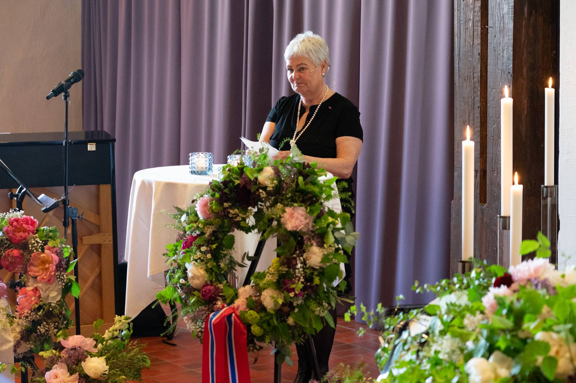 En dame med kort grått hår står ved et bord pyntet med hvit duk og blomster. Hun leser fra kort. 