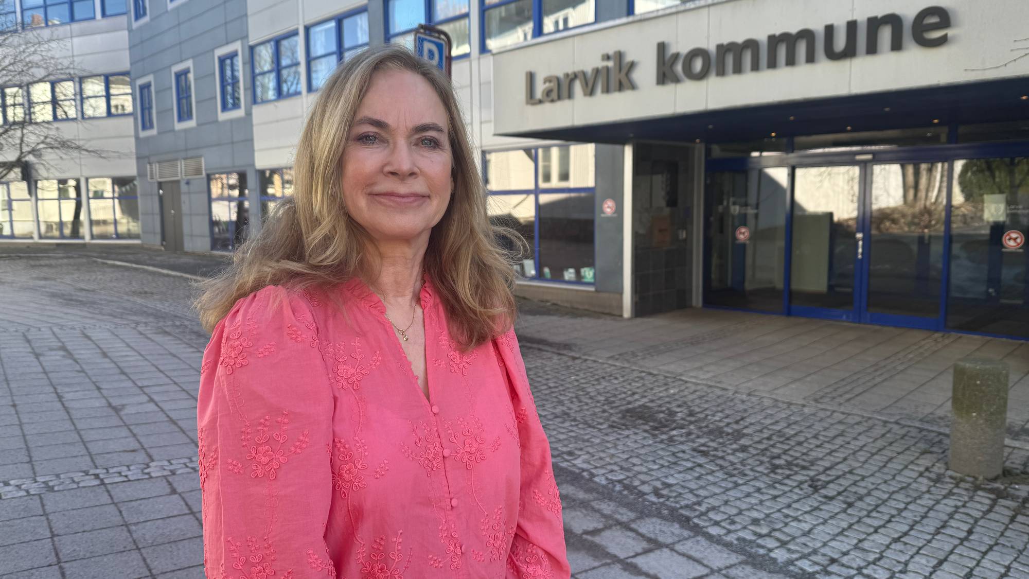En kvinne i rosa bluse som står foran et bygg der det står "Larvik kommune" på fremsiden.