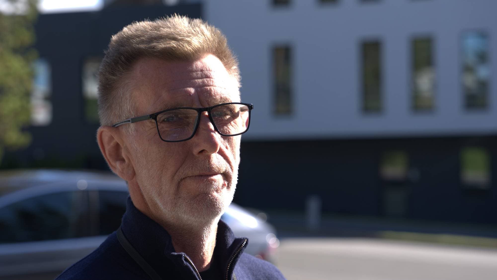 Portrett av en eldre mann med briller og mørk genser utendørs foran en bygning. (Bildebeskrivelsen er laget av en KI-tjeneste)