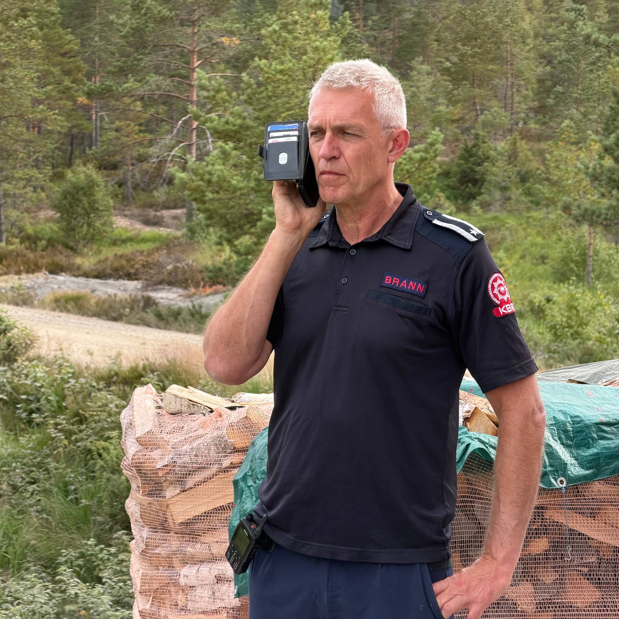Brannkonstabel i uniform snakker i telefon ved en vedstabel i skogsterreng. (Bildebeskrivelsen er laget av en KI-tjeneste)