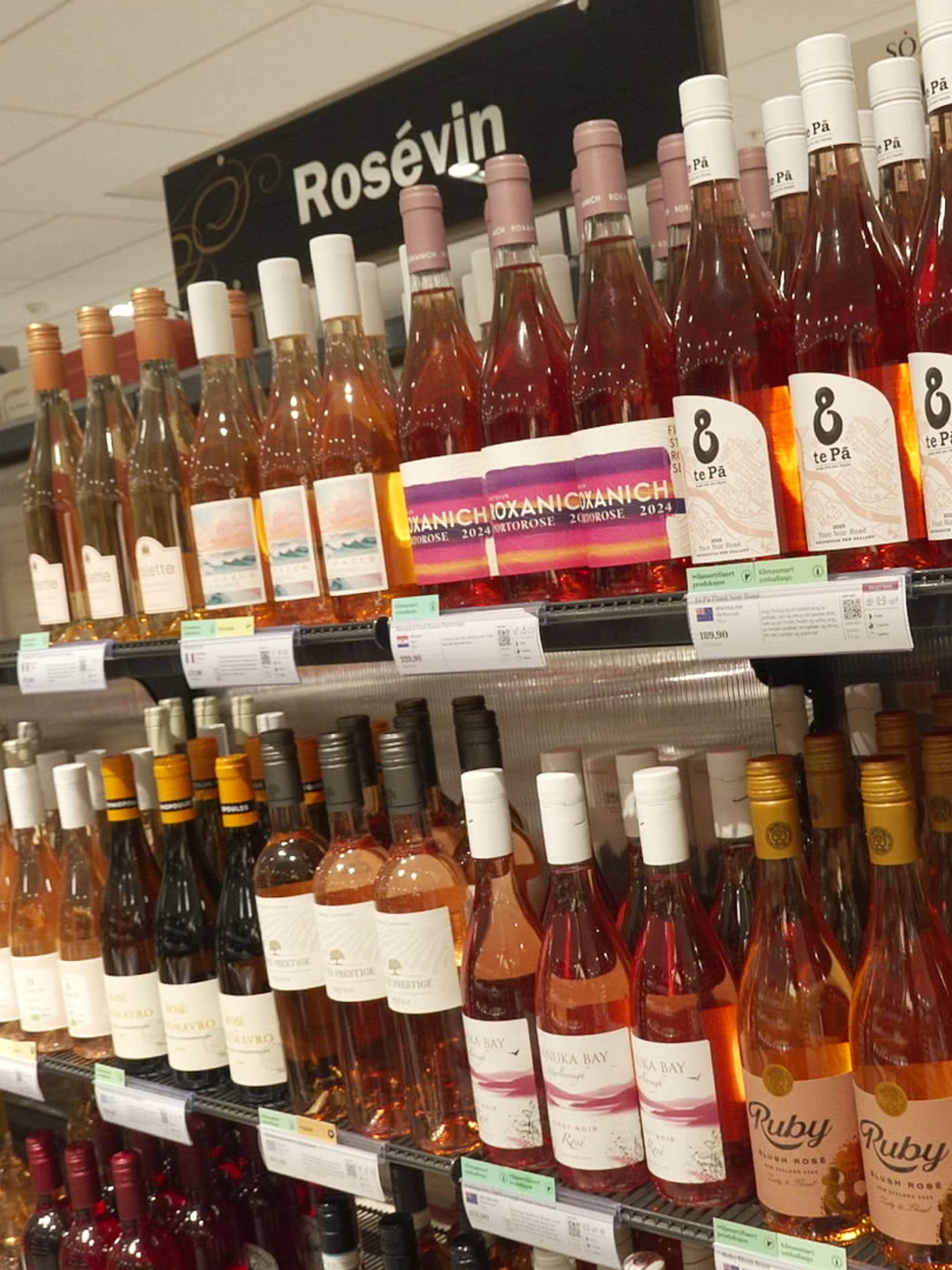 Hyller med rosévin står i en butikkhylle. De fleste flaskene er fylt med rosa eller oransje væske. Noen av flaskene har hvite eller rosa korker, mens andre har mørke korker. Det er prismerker på hyllene. (Bildebeskrivelsen er laget av en KI-tjeneste)