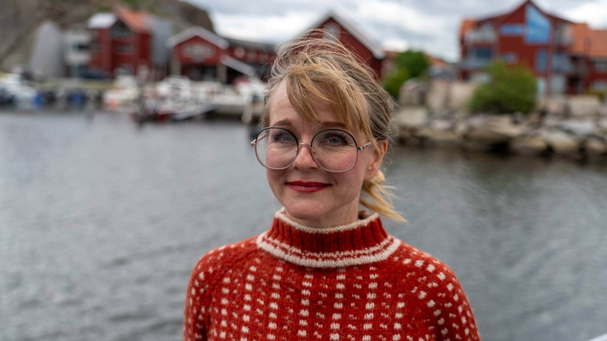 En kvinne med briller og røde lepper smiler mot kameraet. Hun har på seg en rød genser med hvitt mønster. I bakgrunnen ses en brygge med røde trehus og båter. (Bildebeskrivelsen er laget av en KI-tjeneste)