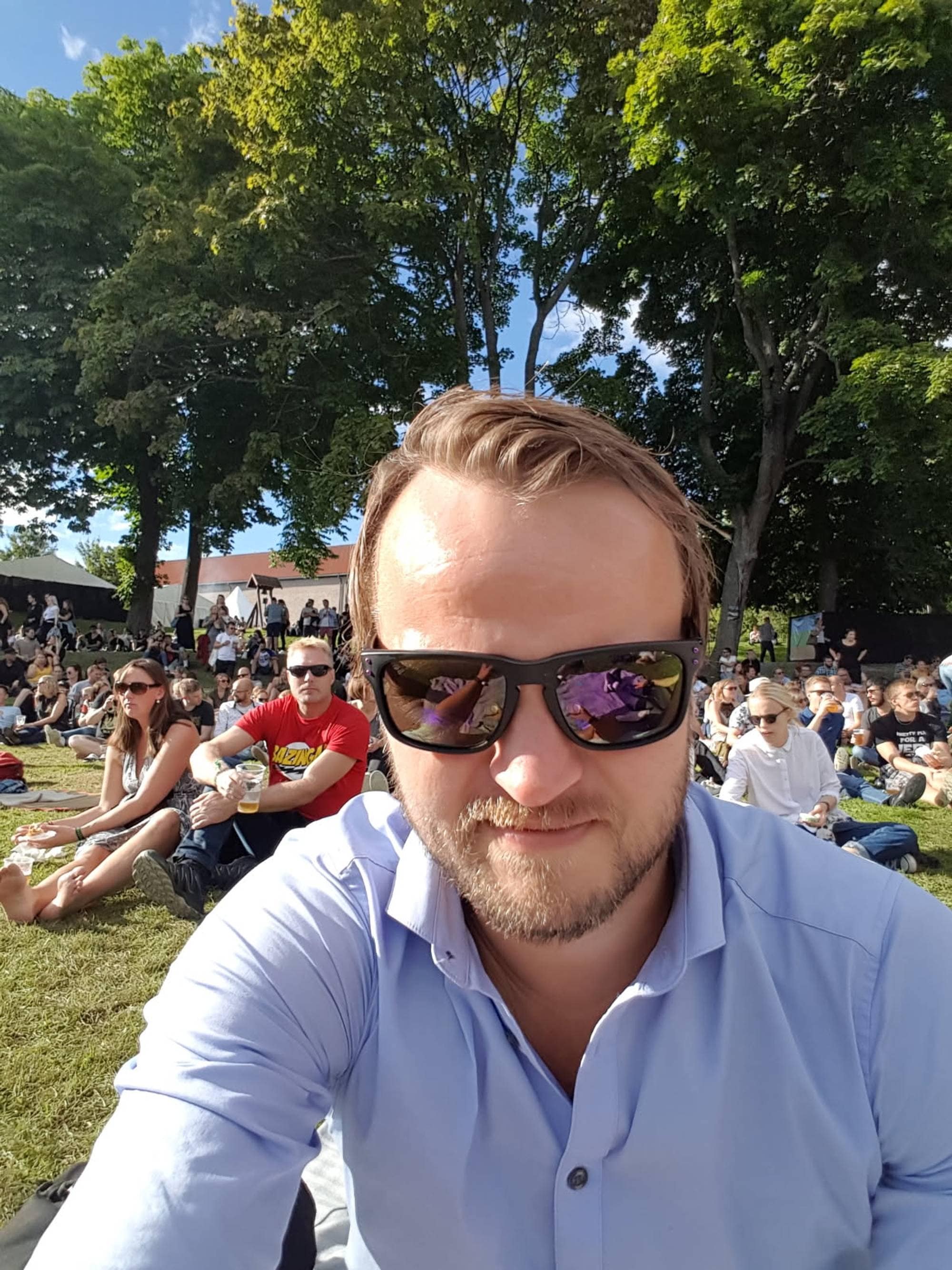 Mann i lyseblå skjorte og solbriller tar bilde av seg selv i en park med publikum i bakgrunnen. (Bildebeskrivelsen er laget av en KI-tjeneste)
