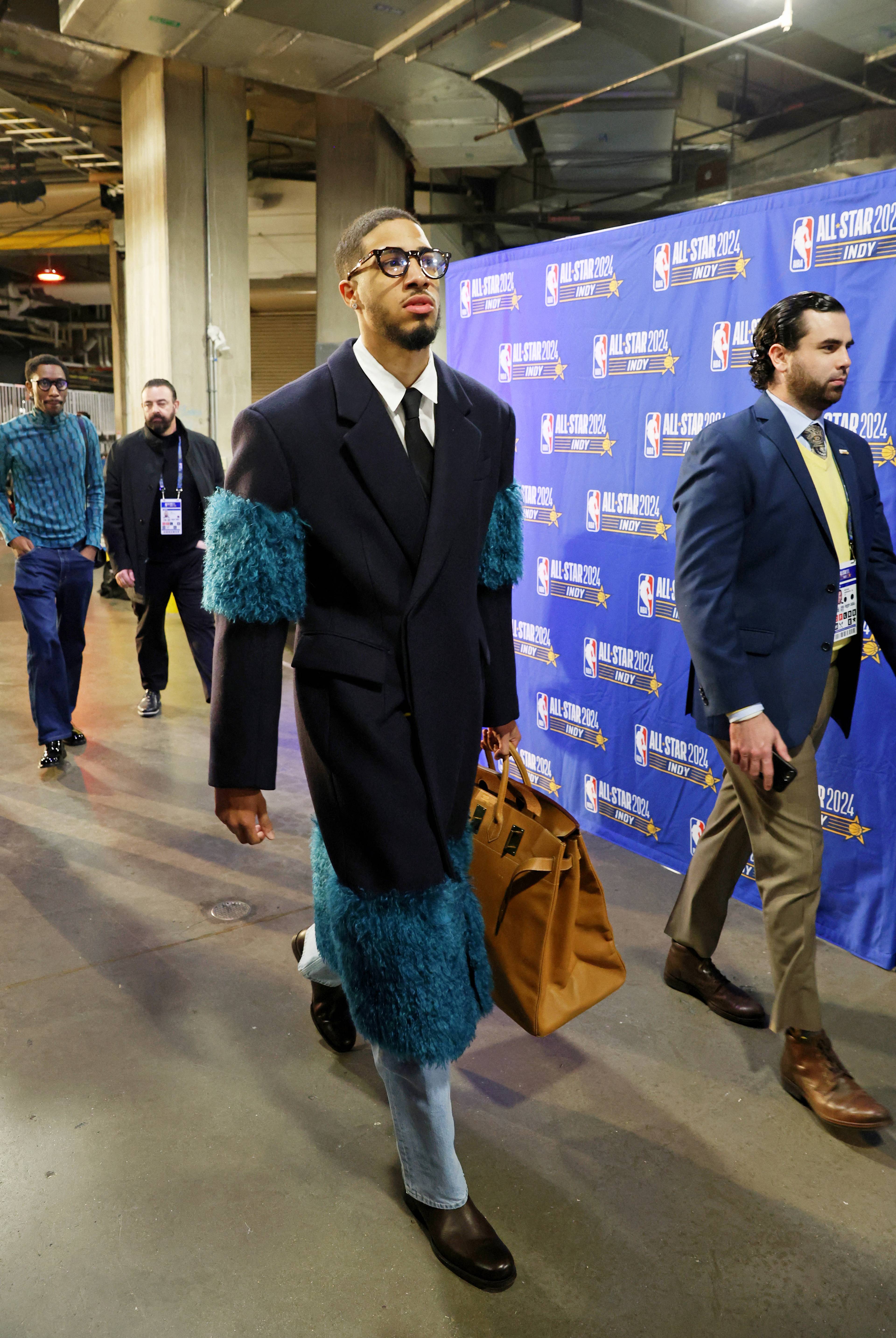 En mann i en stor, mørk frakk med blå pelsdetaljer, jeans og briller går med en veske. Han går forbi en blå bakvegg med logoer for NBA All-Star 2024 Indy. En annen mann i dress følger etter ham. Flere andre personer er også synlige i bakgrunnen. (Bildebeskrivelsen er laget av en KI-tjeneste)