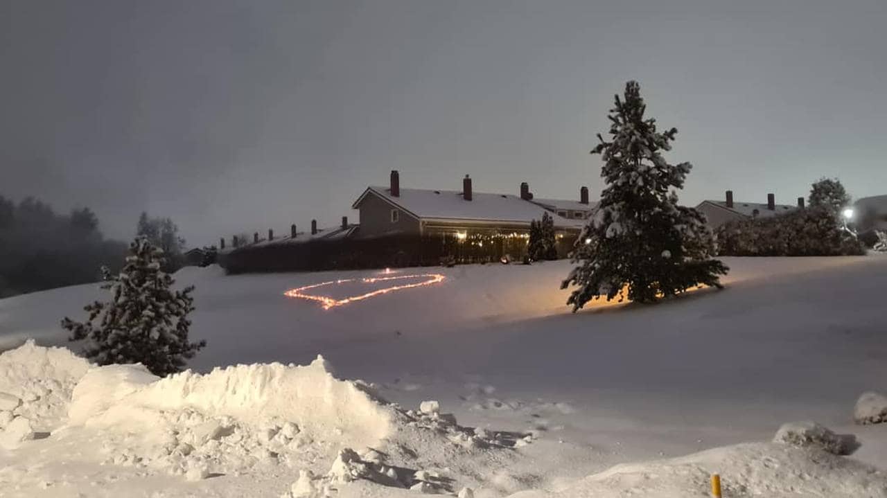 Bilde tatt på avstand av et hjerte i snøen laget av stearinlys. Hjertet hviler på en snødekt skråning opp mot et av husene i Gjerdrum. Det er skumring. 
