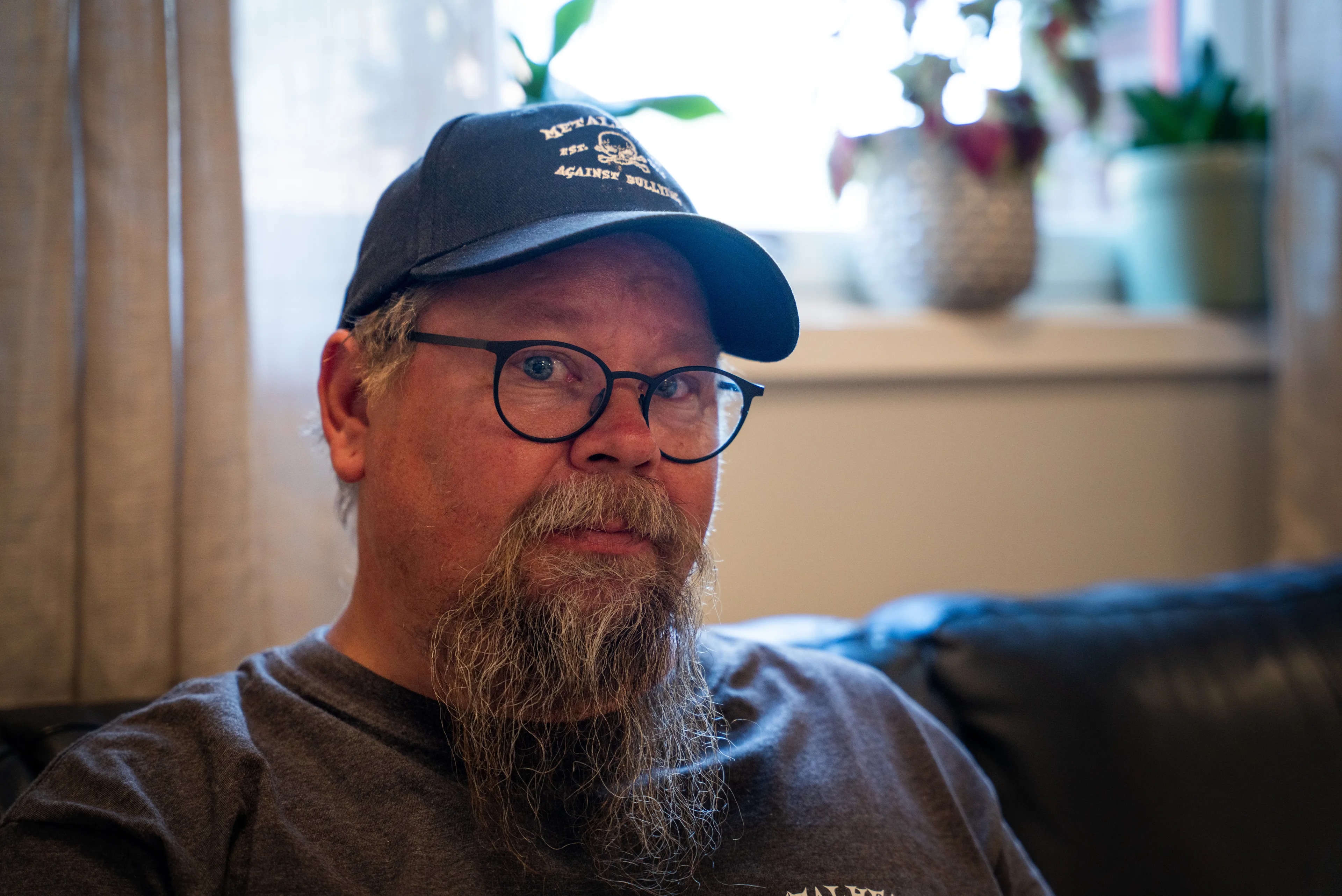 En mann med skjegg, briller og caps sitter inne ved et vindu. (Bildebeskrivelsen er laget av en KI-tjeneste)