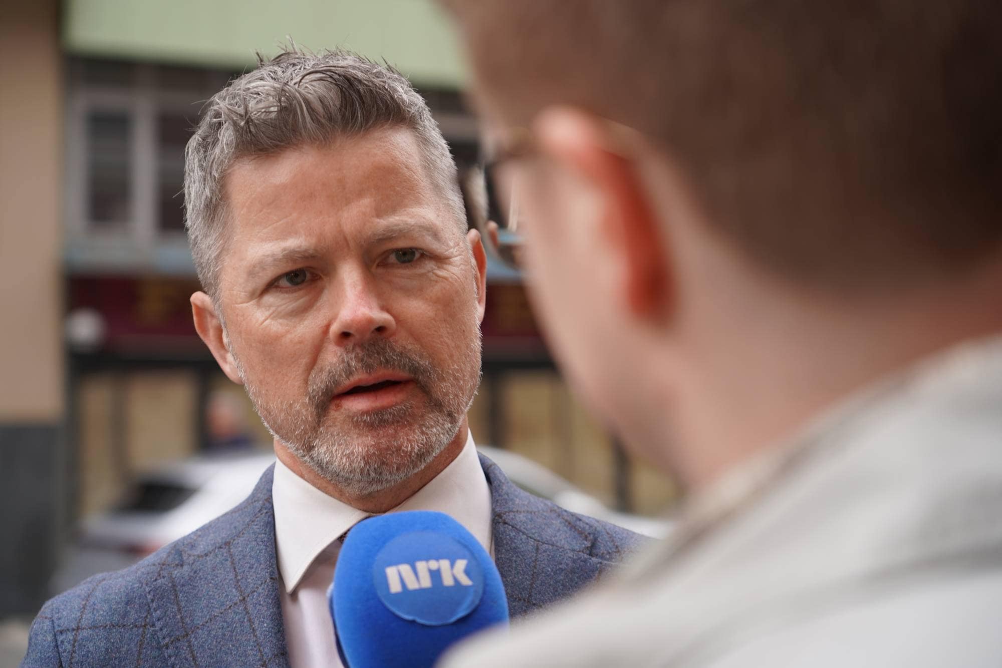 En mann med grått skjegg og dress blir intervjuet av en person ute. Mannen holder en mikrofon med NRK-logo. Det er en bygate i bakgrunnen. (Bildebeskrivelsen er laget av en KI-tjeneste)