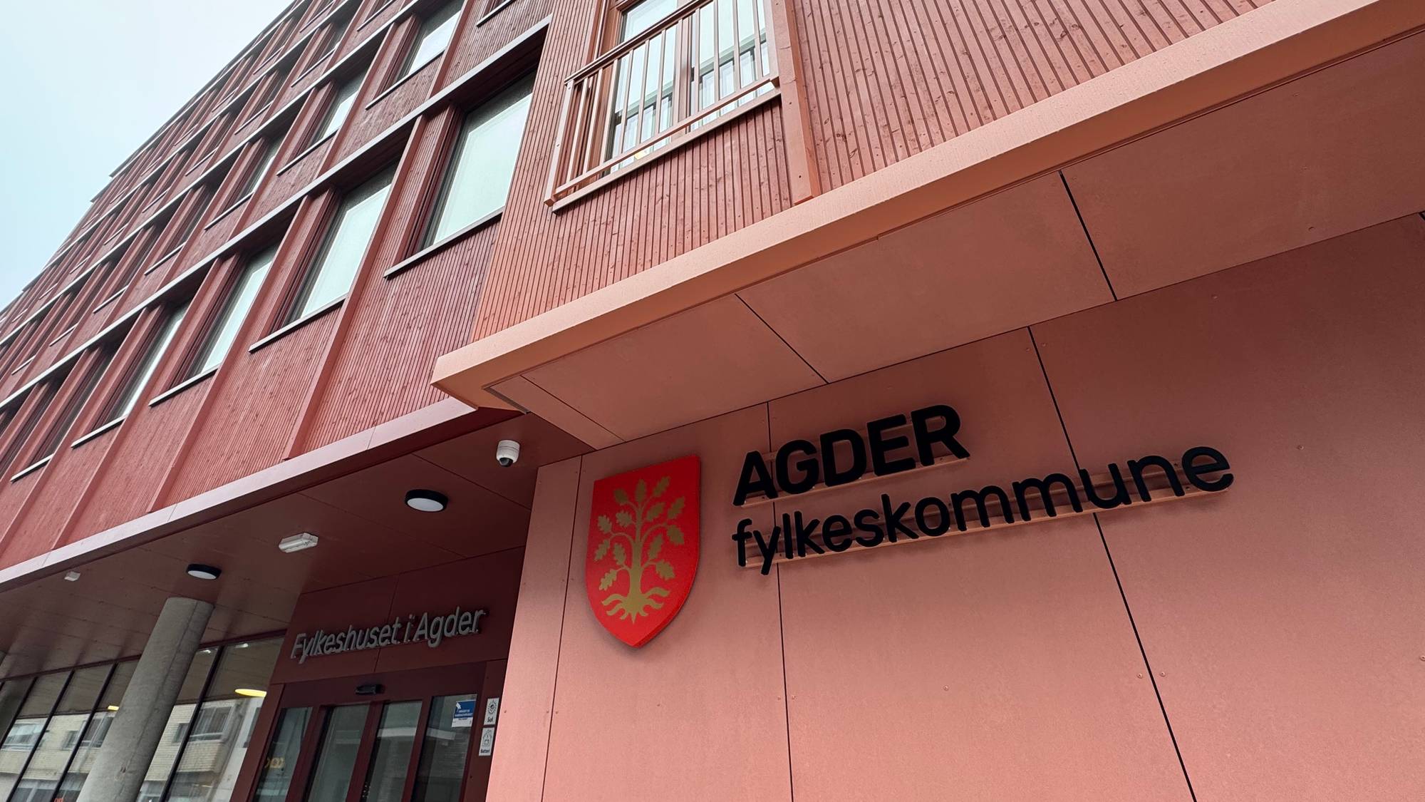 Fylkeshuset i Agder fotografert fra bakkenivå med fokus på bygningens fasade. Bygget har røde vegger og flere vinduer. Skiltet med teksten "AGDER fylkeskommune" er synlig ved inngangen. Omgivelsene er grå og skyet, noe som gir et dyster preg. (Bildebeskrivelsen er laget av en KI-tjeneste)