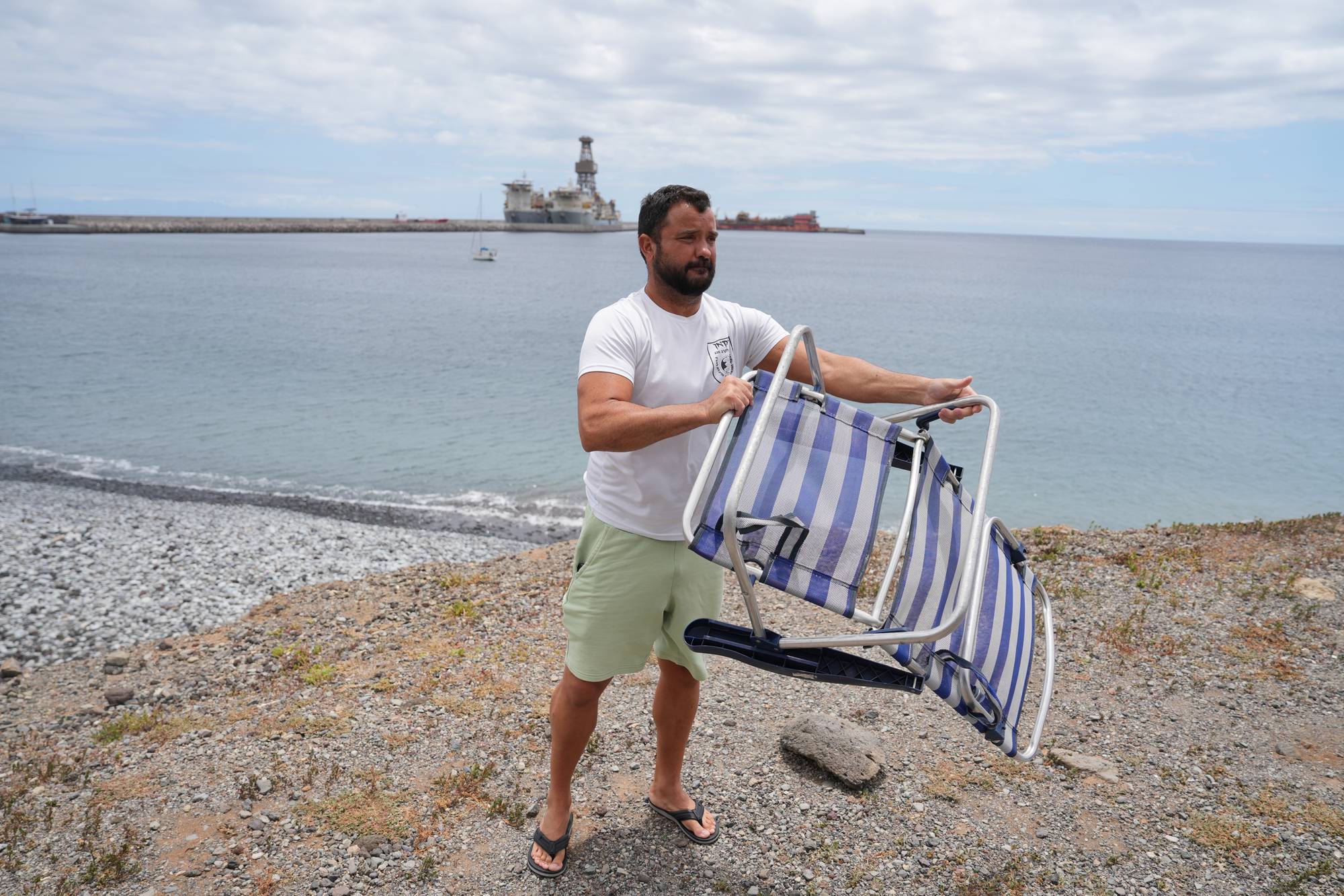 En mann med skjegg bretter ut en campingstol. Han har på seg hvit t-skjorte og lysegrønn shorts. Bak ham er det en strand, hav og en industriell installasjon på vannet. 
