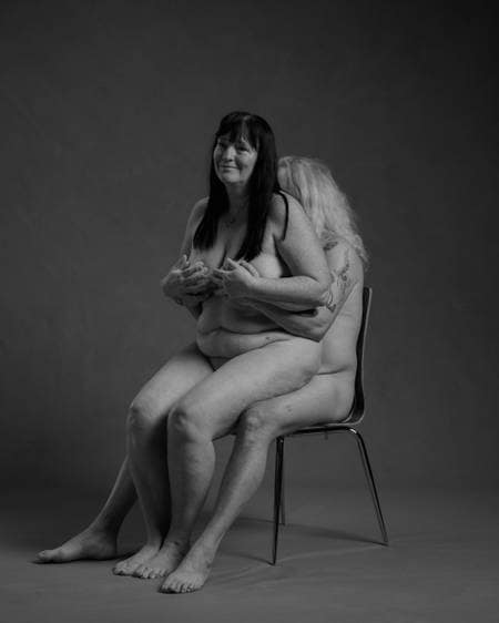 En naken dame med langt mørkt hår sitter på fanget til en naken mann med langt hvitt hår som sitter på en stol og holder henne på puppene. De er et godt voksent par.