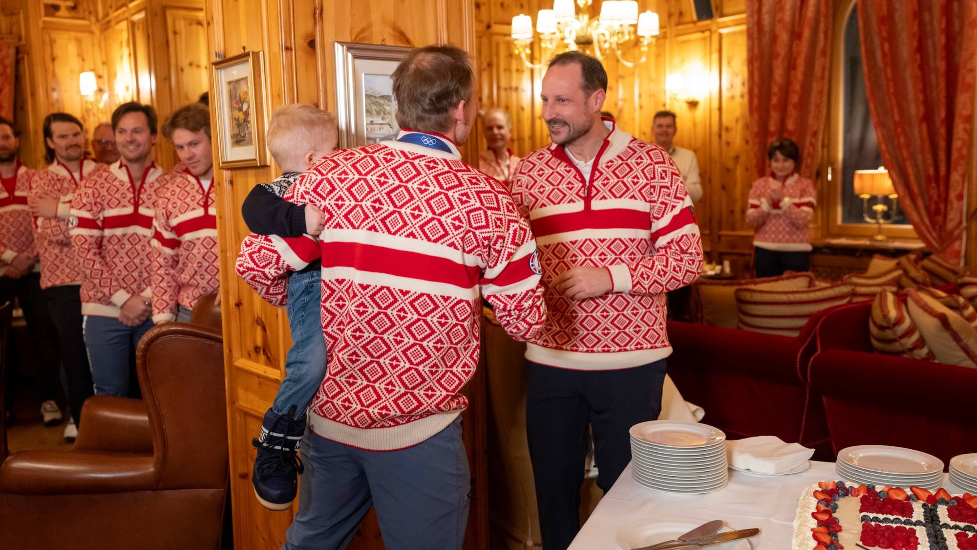 Kronprins Haakon hilser på en mann som holder et barn, begge iført norske OL-gensere. (Bildebeskrivelsen er laget av en KI-tjeneste)