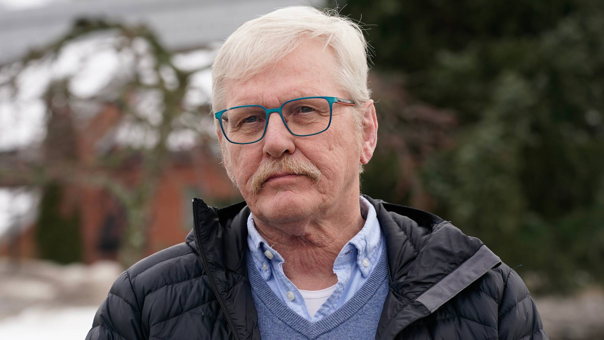 Portrett av en eldre mann med briller og bart, kledd i blå genser og svart jakke. (Bildebeskrivelsen er laget av en KI-tjeneste)