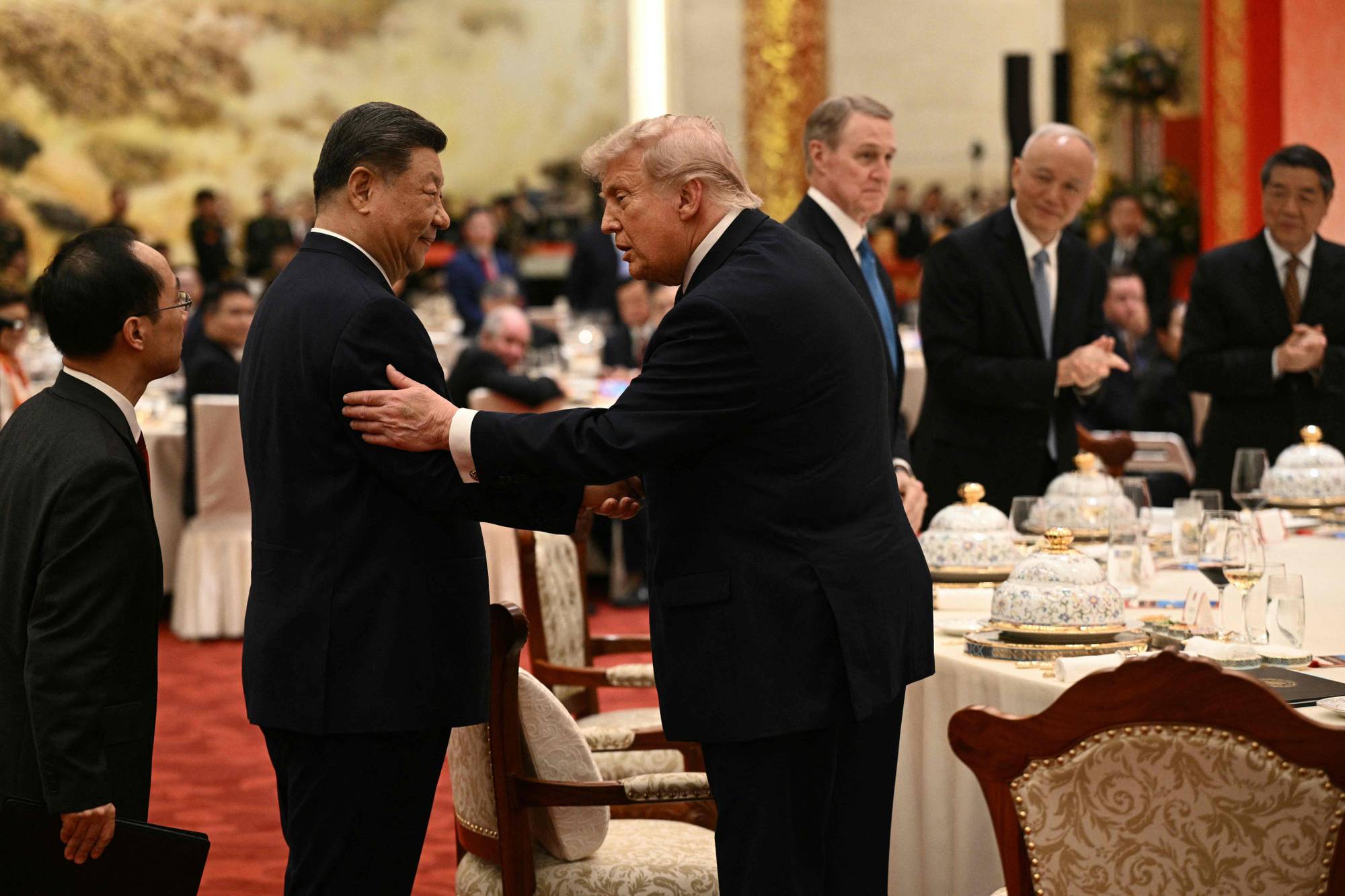 Kinas president Xi Jinping og USAs president Donald Trump hilser på hverandre ved et bord. Flere andre menn i dress er synlige i bakgrunnen. De sitter ved et langbord dekket med servise. (Bildebeskrivelsen er laget av en KI-tjeneste)