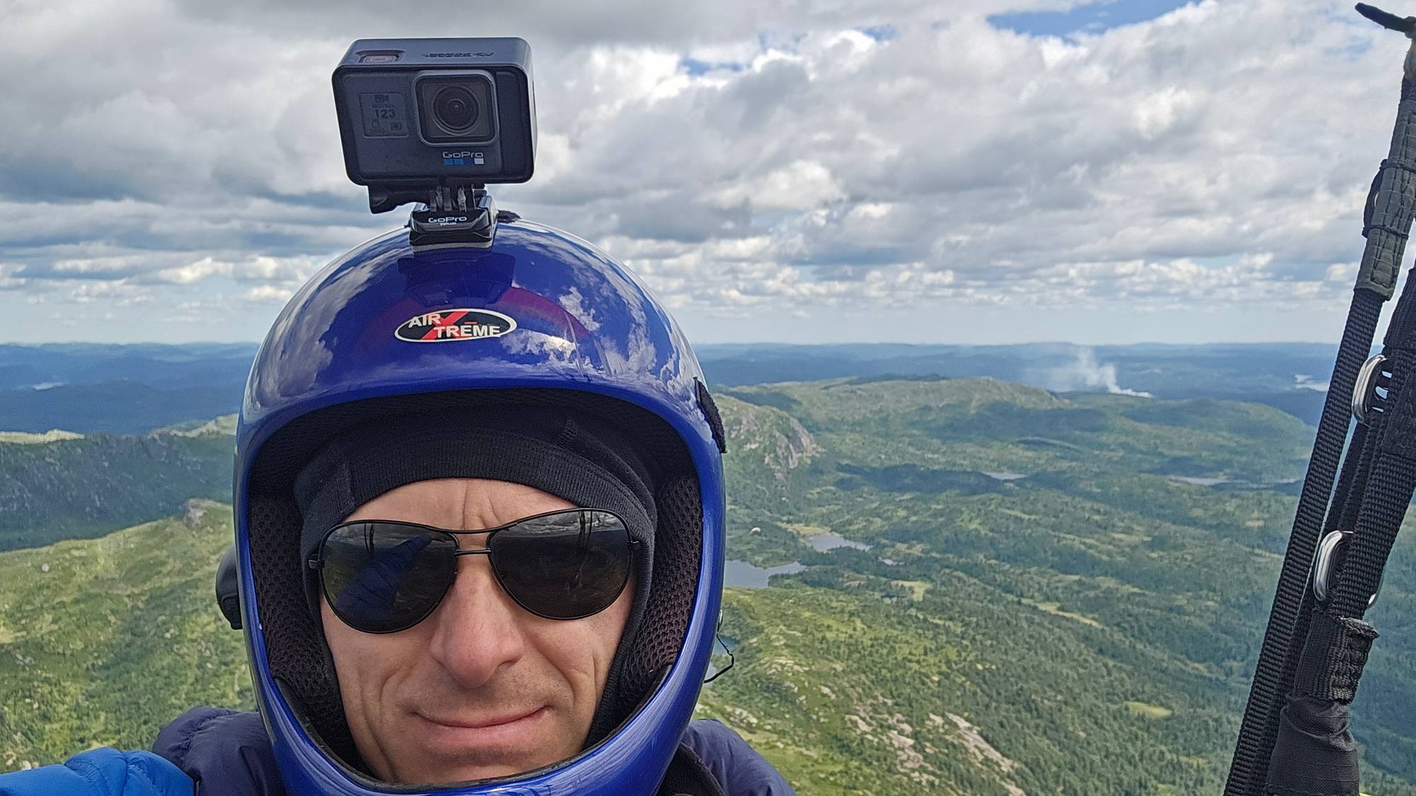 Person med hjelm og kamera i paraglider over et landskap med fjell og vann. (Bildebeskrivelsen er laget av en KI-tjeneste)