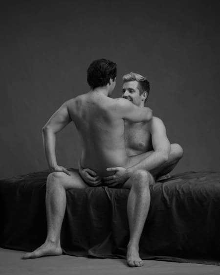 To nakne menn på en mørk seng. Mannen med lyst hår sitter på sengekanten. Mannen med mørkt hår sitter på fanget til partneren med beina rundt midjen hans. Han har venstre hånd på den andre mannens kne og høyre hånd rundt halsen hans. De ser på hverandre. Mannen med lyst hår har hendene på rumpa til den andre.