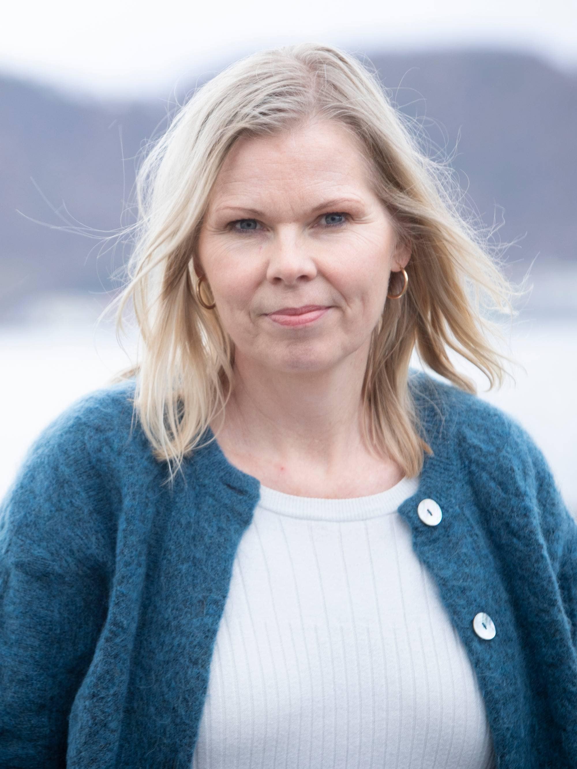 En kvinne i blå cardigan og hvit tskjorte og lyst halvlangt hår står utendørs og ser inn i kamera