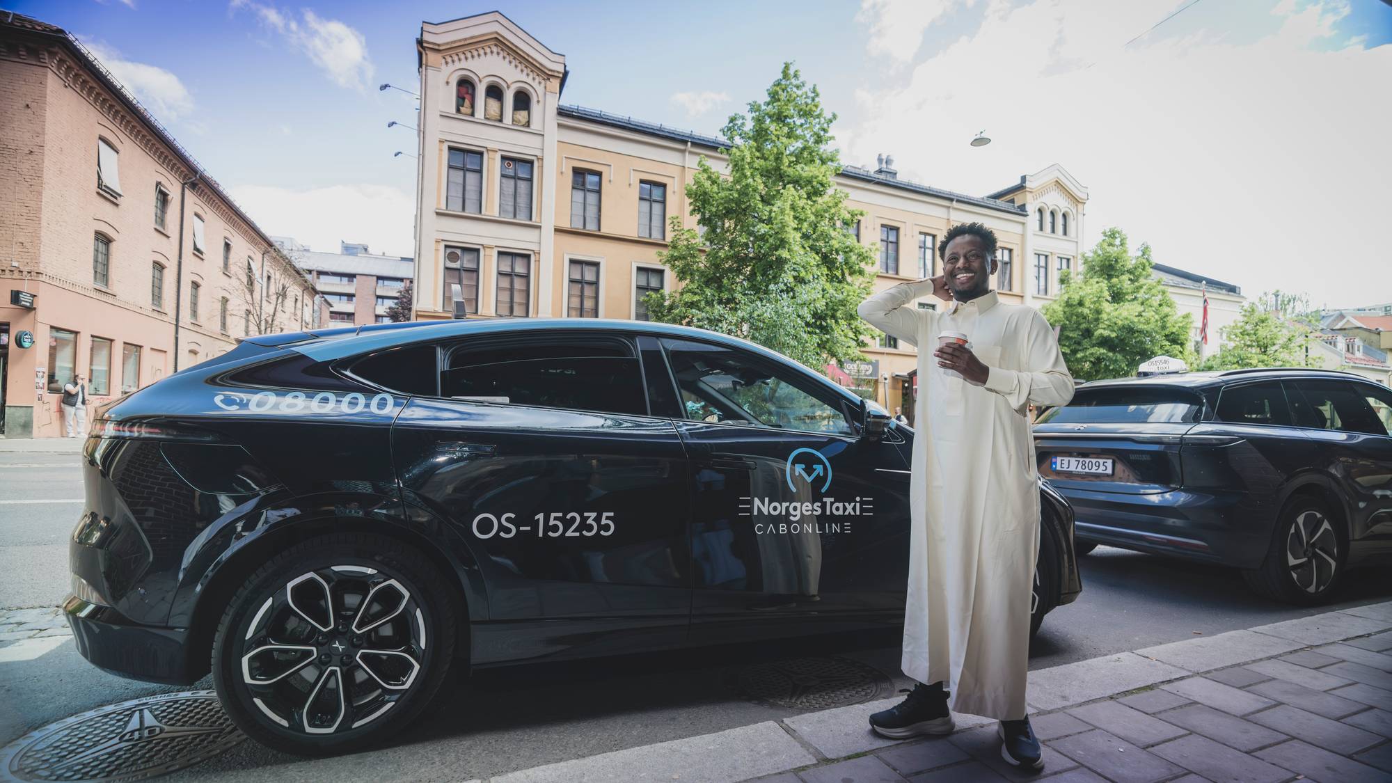 En blid mann holder en kaffekopp og står foran bilen sin.