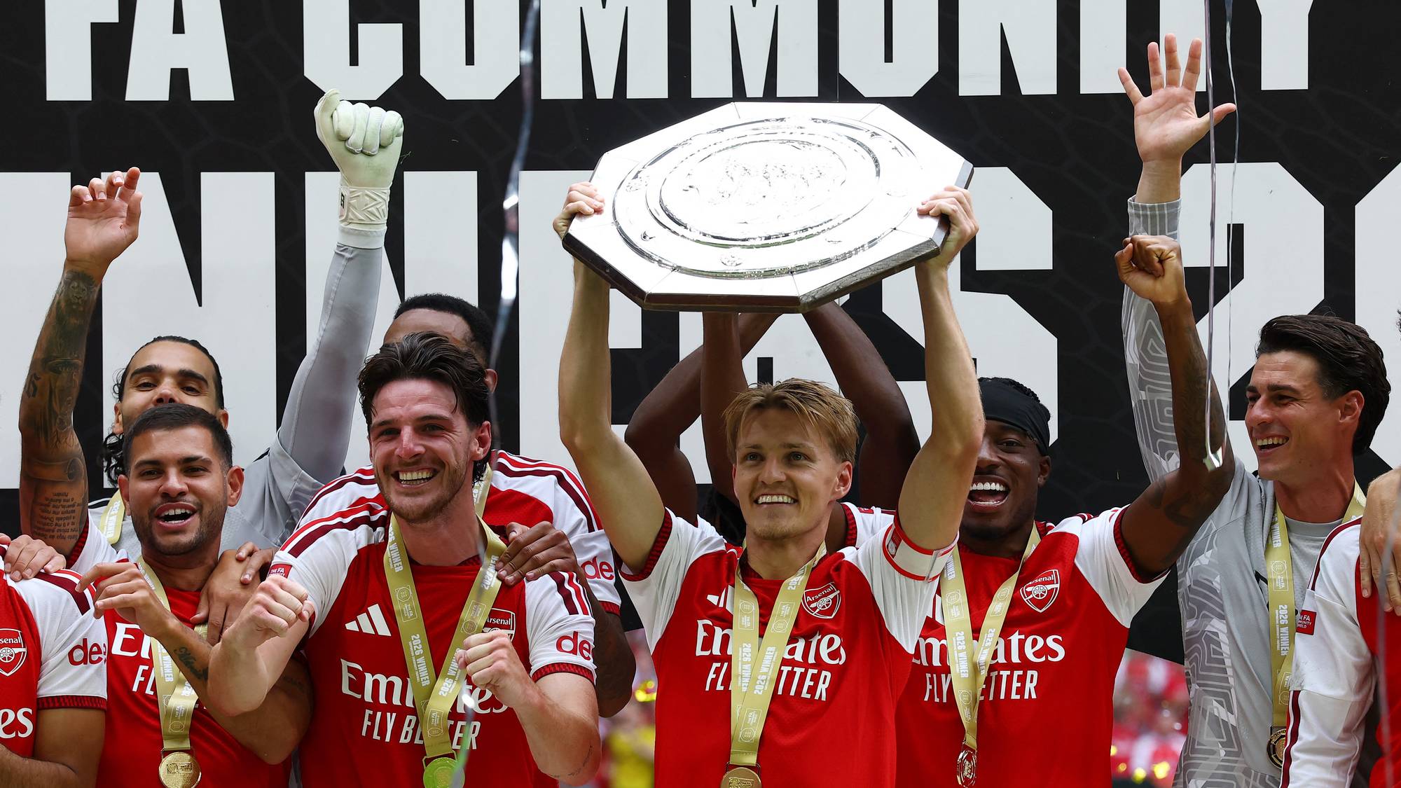 Arsenal-spillere feirer seier i FA Community Shield med pokalen hevet foran en plakat. (Bildebeskrivelsen er laget av en KI-tjeneste)