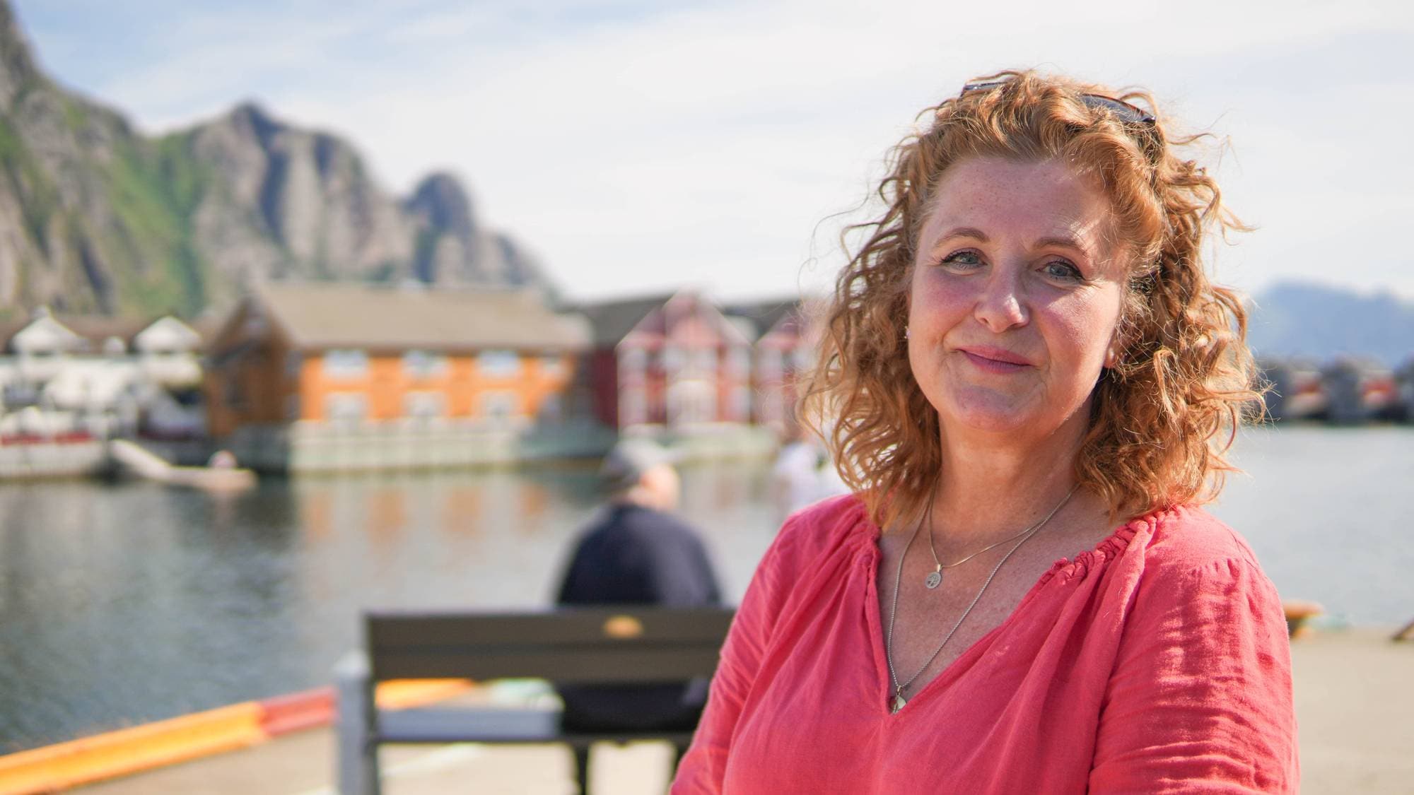 Line Samuelsen, reiselivssjef i Lofoten fotografert på kaia i Svolvær. Hun har rødt krøllete hår og en rosa topp på seg. I bakgrunnen sitter en turist på en benk, og fjellet brer seg over landskapet.