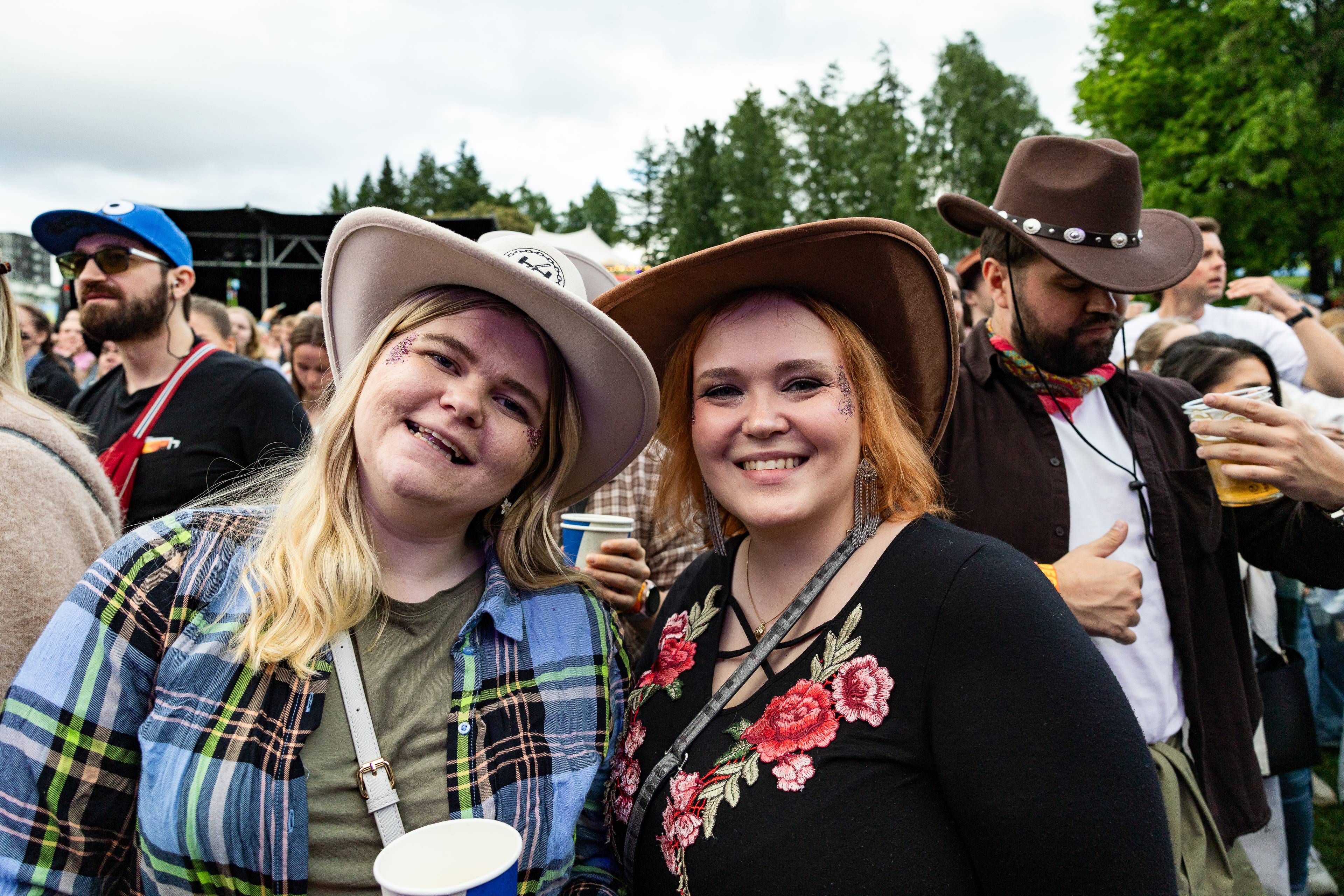 To kvinner i cowboyhatter smiler til kameraet, med et publikum bak.Begge kvinnene har glitter i ansiktet.En mann i bakgrunnen med en brun cowboyhatt holder en øl.Bildet er tatt utendørs med trær og en scene i bakgrunnen. (Bildebeskrivelsen er laget av en KI-tjeneste)