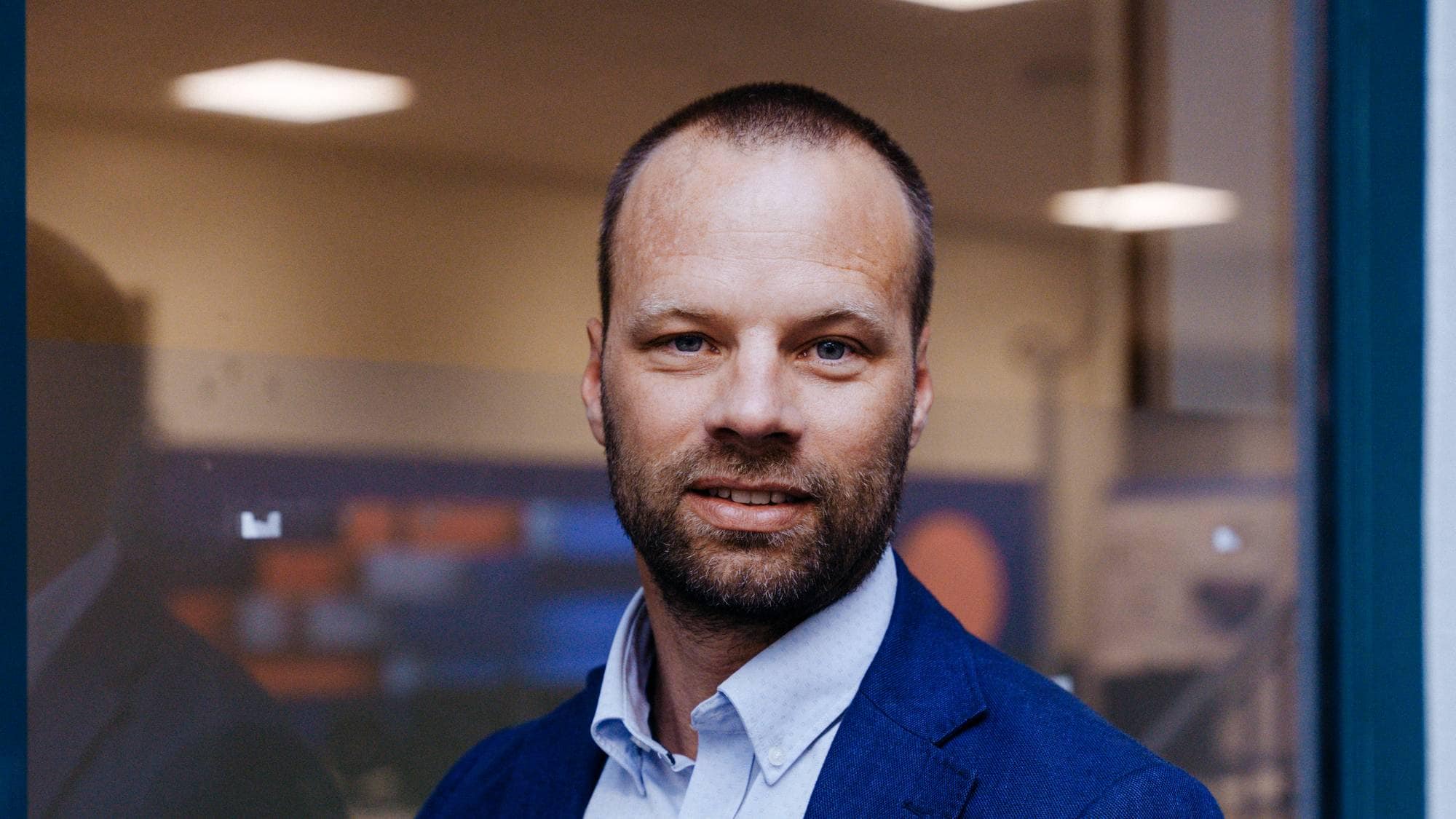 Portrett av en mann i blå dressjakke og lys skjorte, fotografert foran en glassvegg. 