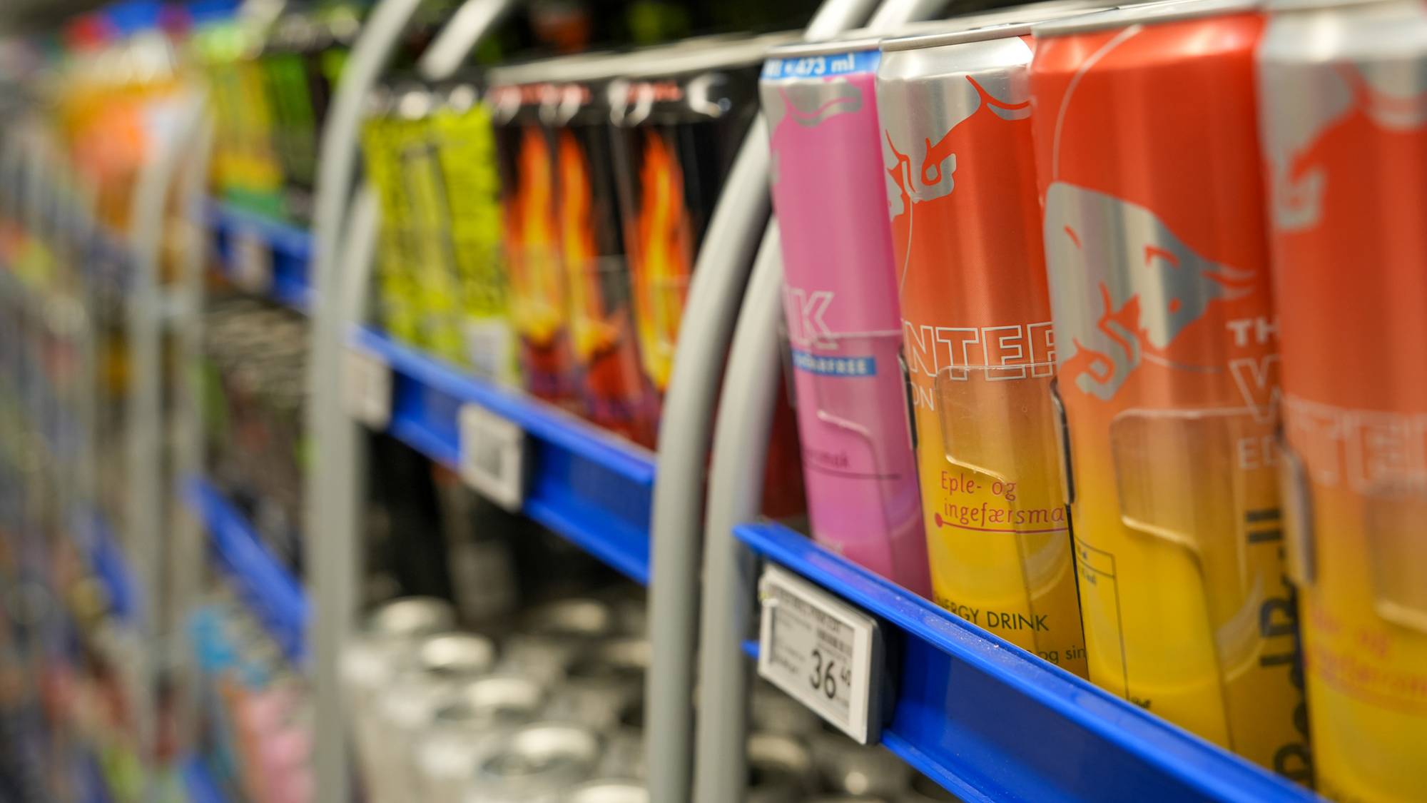 Bildet viser en rekke energidrikker plassert på hyller i en butikk. Det er flere ulike farger og design på boksene, inkludert merker som Red Bull. Høyre siden viser bokser med oransje, lyserosa og gule farger med påskrift. Bakgrunnen er uskarp, hvilket fokuserer oppmerksomheten på drikkene.