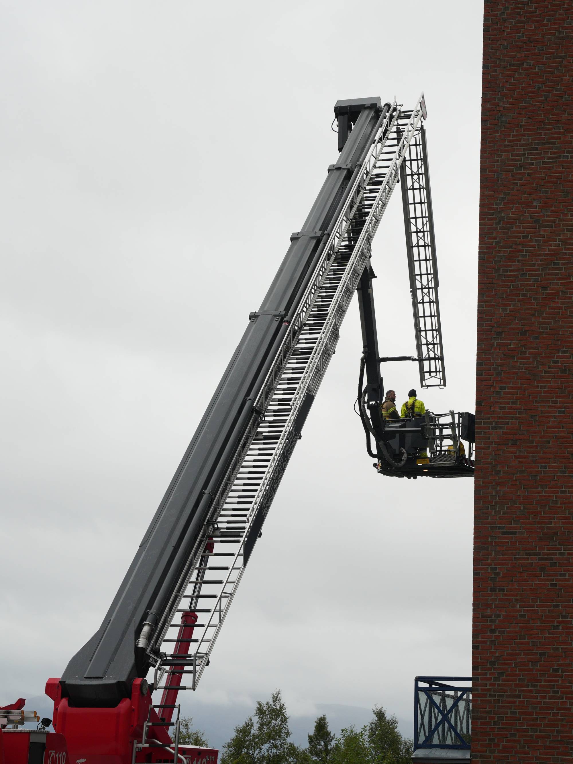 Brannmannskaper i heis på en lang stigebil ved en rød murvegg. (Bildebeskrivelsen er laget av en KI-tjeneste)