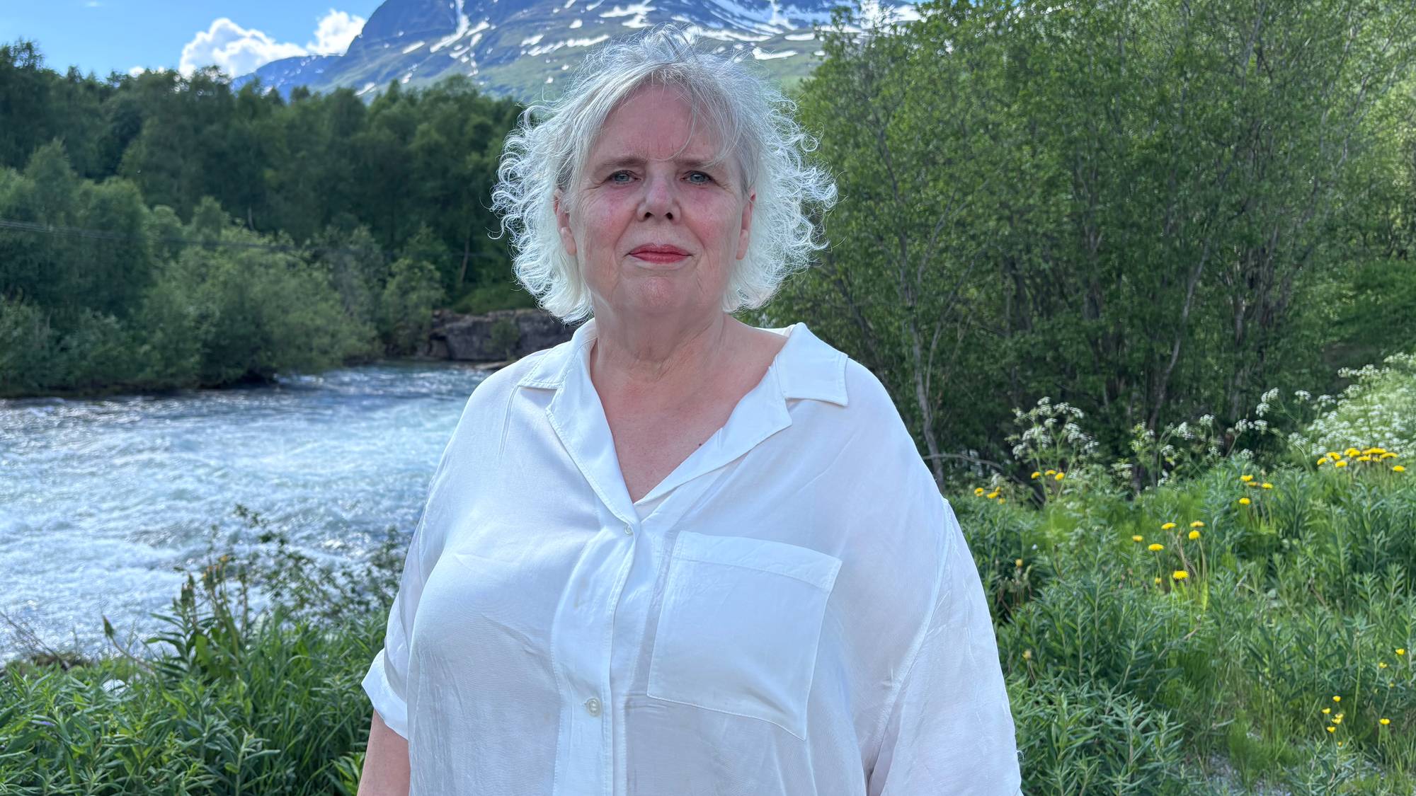 En kvinne står foran en elv med fjell i bakgrunnen. Hun har grått hår og har på seg en hvit skjorte. Elven renner gjennom et grønt landskap med trær og busker. (Bildebeskrivelsen er laget av en KI-tjeneste)
