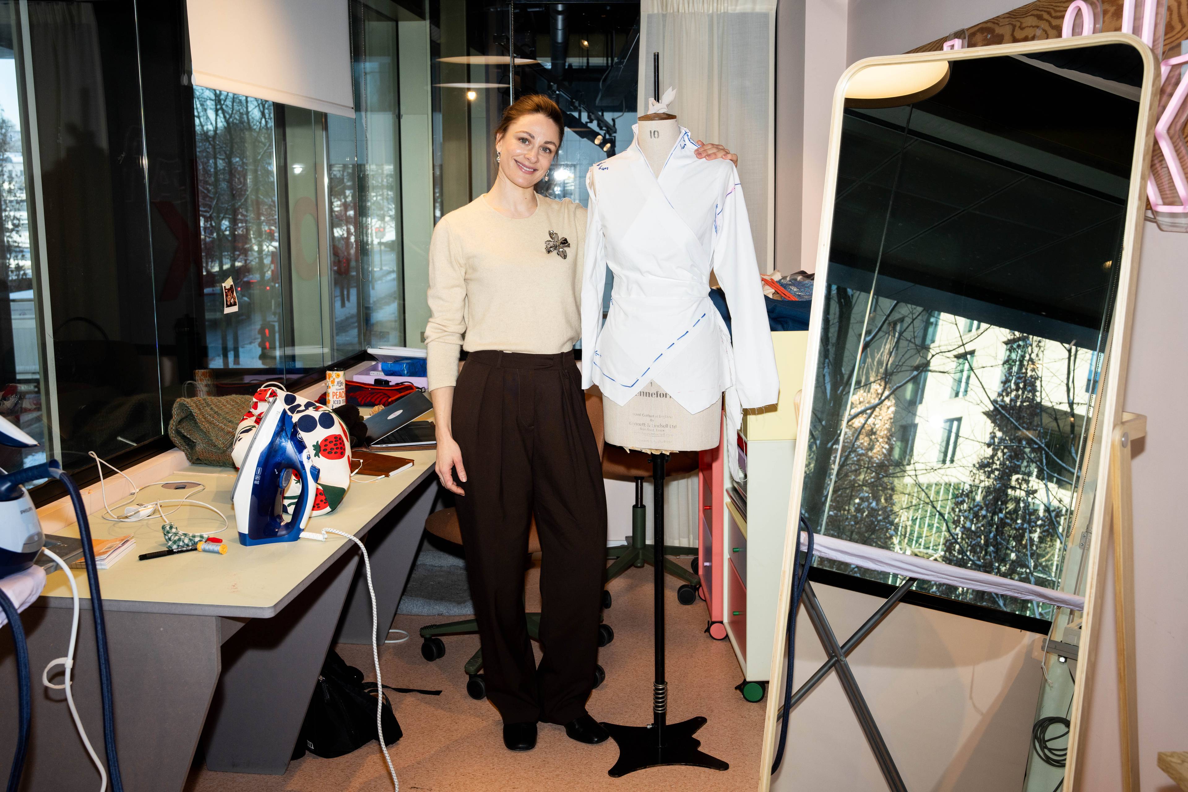 Jenny Skavlan fotografert i et syrom. Hun står ved siden av et mannekeng og rundt henne er syverktøy på en benk og et speil.