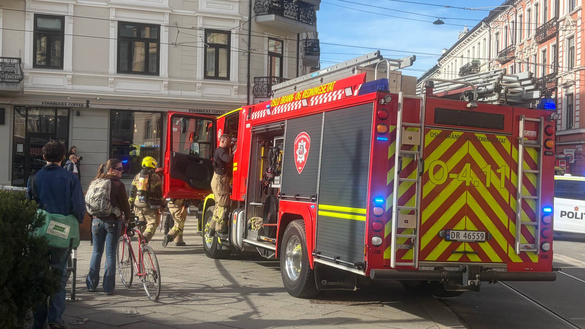 Brannbil fra Oslo brann- og redningsetat er synlig på en gate i byen. Flere personer, deriblant brannfolk i uniform, er til stede ved siden av kjøretøyet. I bakgrunnen sees bygninger og en politibil. En person holder en sykkel. (Bildebeskrivelsen er laget av en KI-tjeneste)