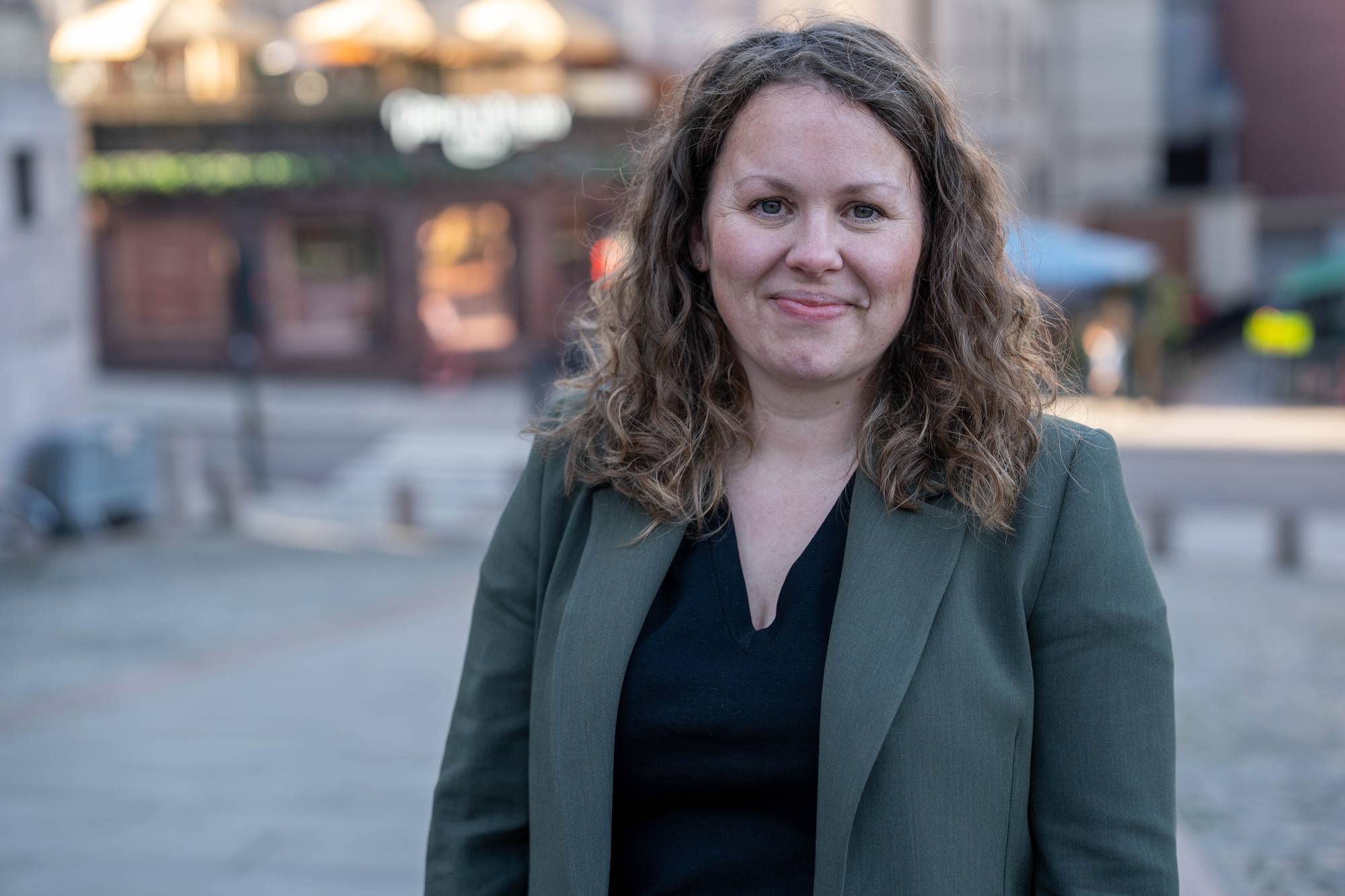 En kvinne står utendørs foran en utydelig bygning. Hun har på seg en mørk jakke over en mørk topp. Kvinnen har krøllete brunt hår og smiler lett. Bak henne skimtes en bygate med forretninger og lyktestolper i en uskarp bakgrunn. (Bildebeskrivelsen er laget av en KI-tjeneste)