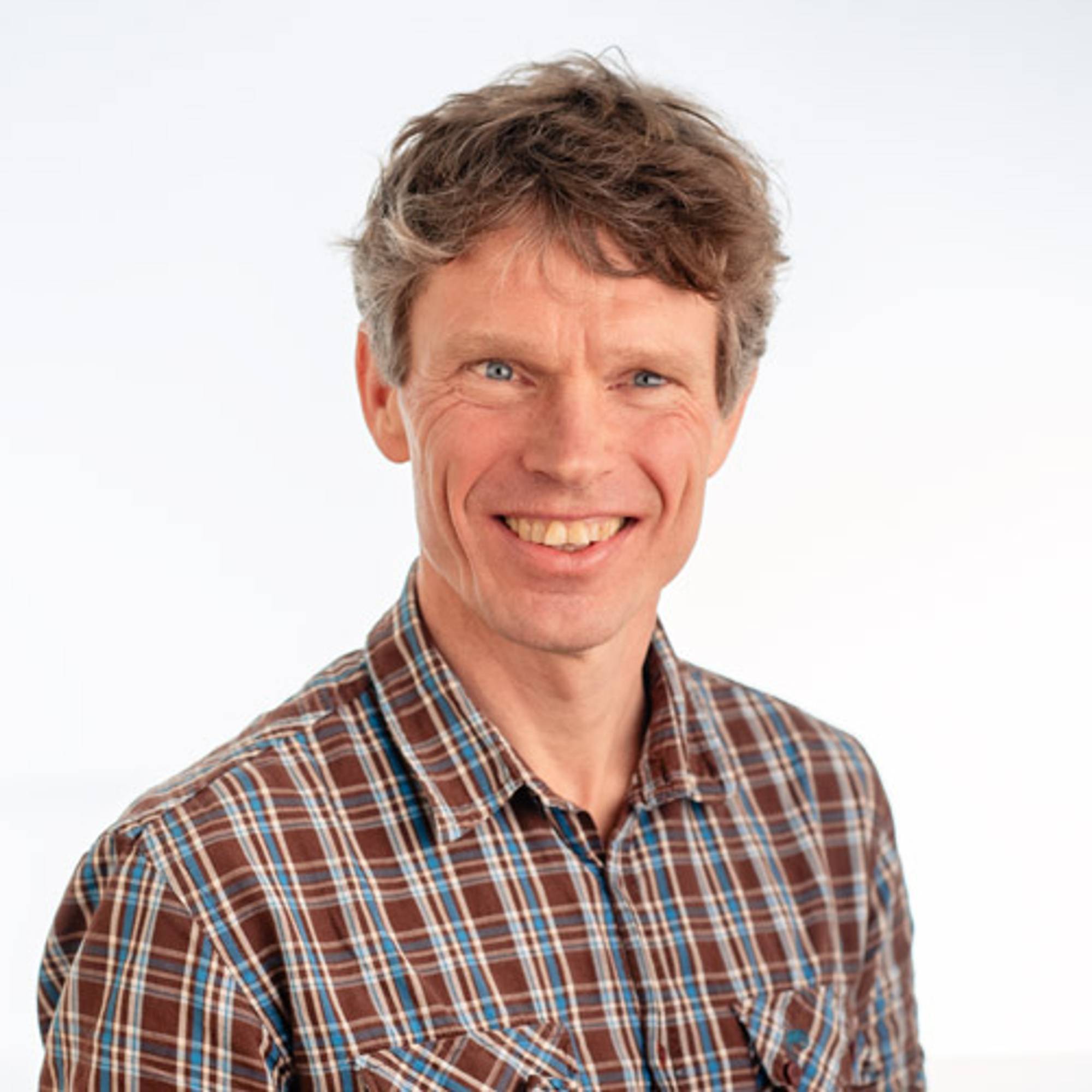 Bildet viser en mann med lysere hår iført en rutete skjorte med flere farger. Han står i et lyst rom med en enkel bakgrunn. Mannen retter blikket mot kameraet, og ansiktsuttrykket hans er nøytral. Det er naturlig lys i rommet, noe som gir en hyggelig atmosfære. (Bildebeskrivelsen er laget av en KI-tjeneste)