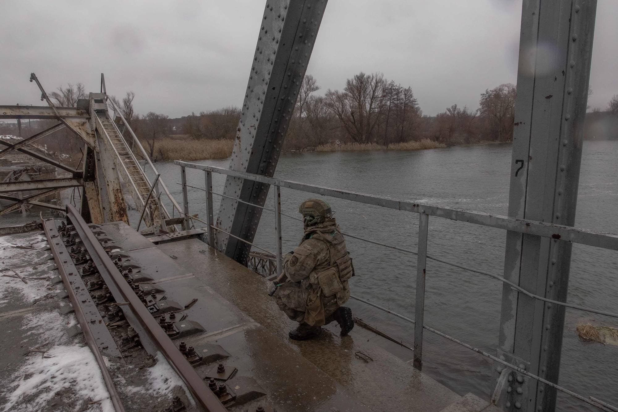 En soldat i kamuflasjedrakt sitter på huk på en metallbro. Broen har jernbanespor og en stige som leder oppover. Ved siden av soldaten renner en elv under en grå himmel. (Bildebeskrivelsen er laget av en KI-tjeneste)