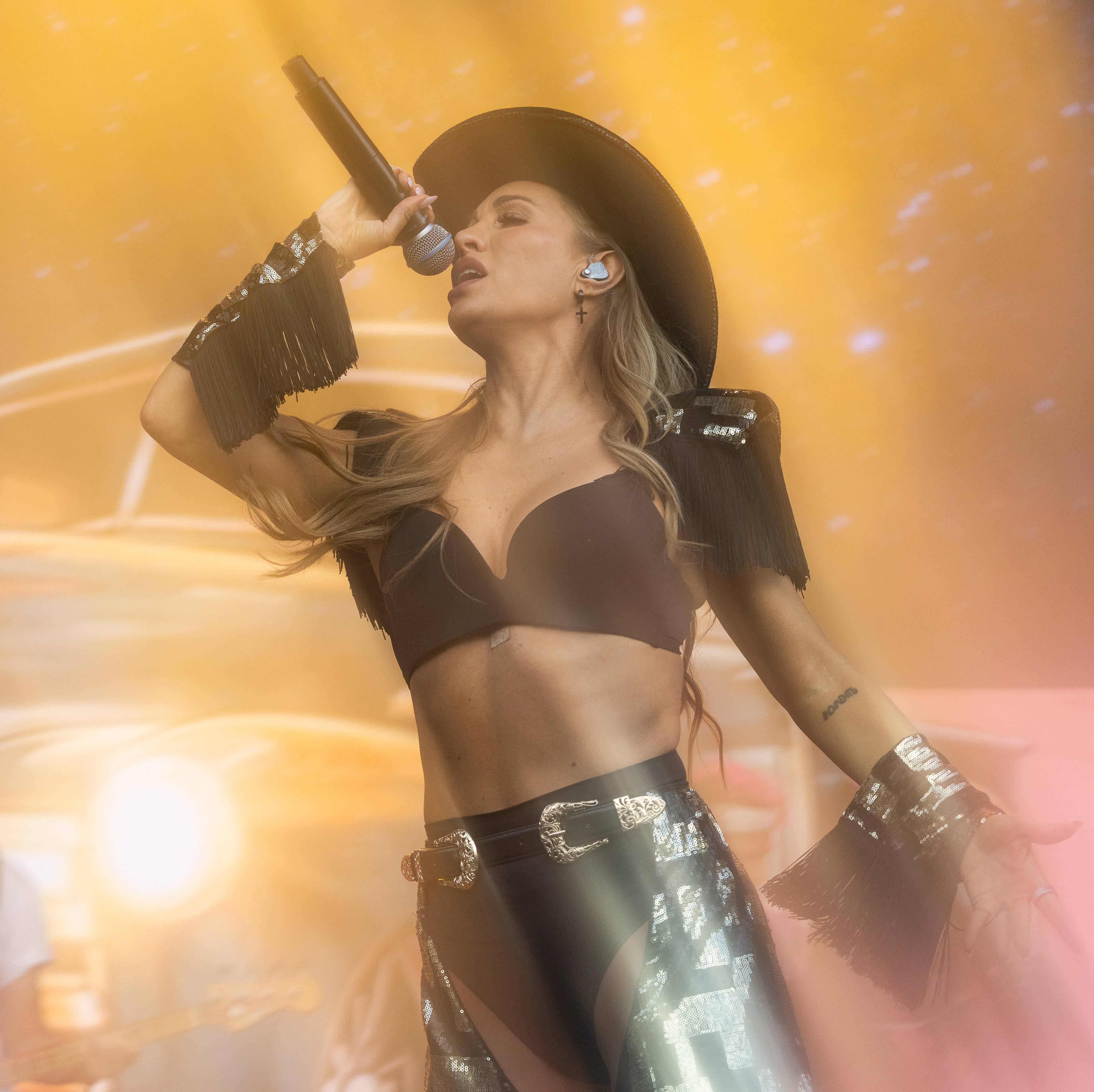 En kvinne i cowboyhatt og mørk topp synger i en mikrofon. Hun har på seg en glitrende bukse og armbånd med frynser. Bak henne sitter en mann med gitar. (Bildebeskrivelsen er laget av en KI-tjeneste)