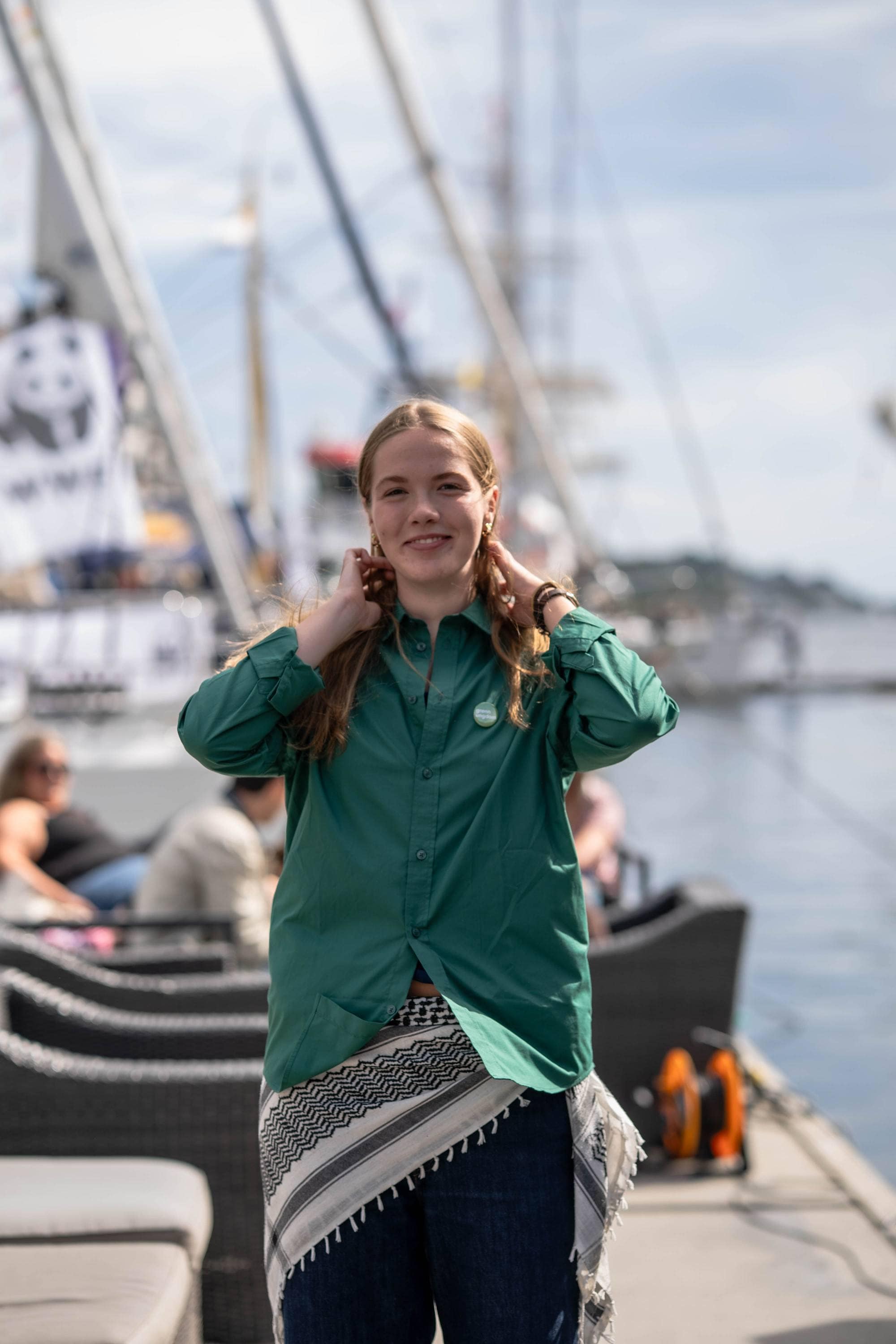 Kvinne i grønn skjorte står på en kai med seilskuter i bakgrunnen.