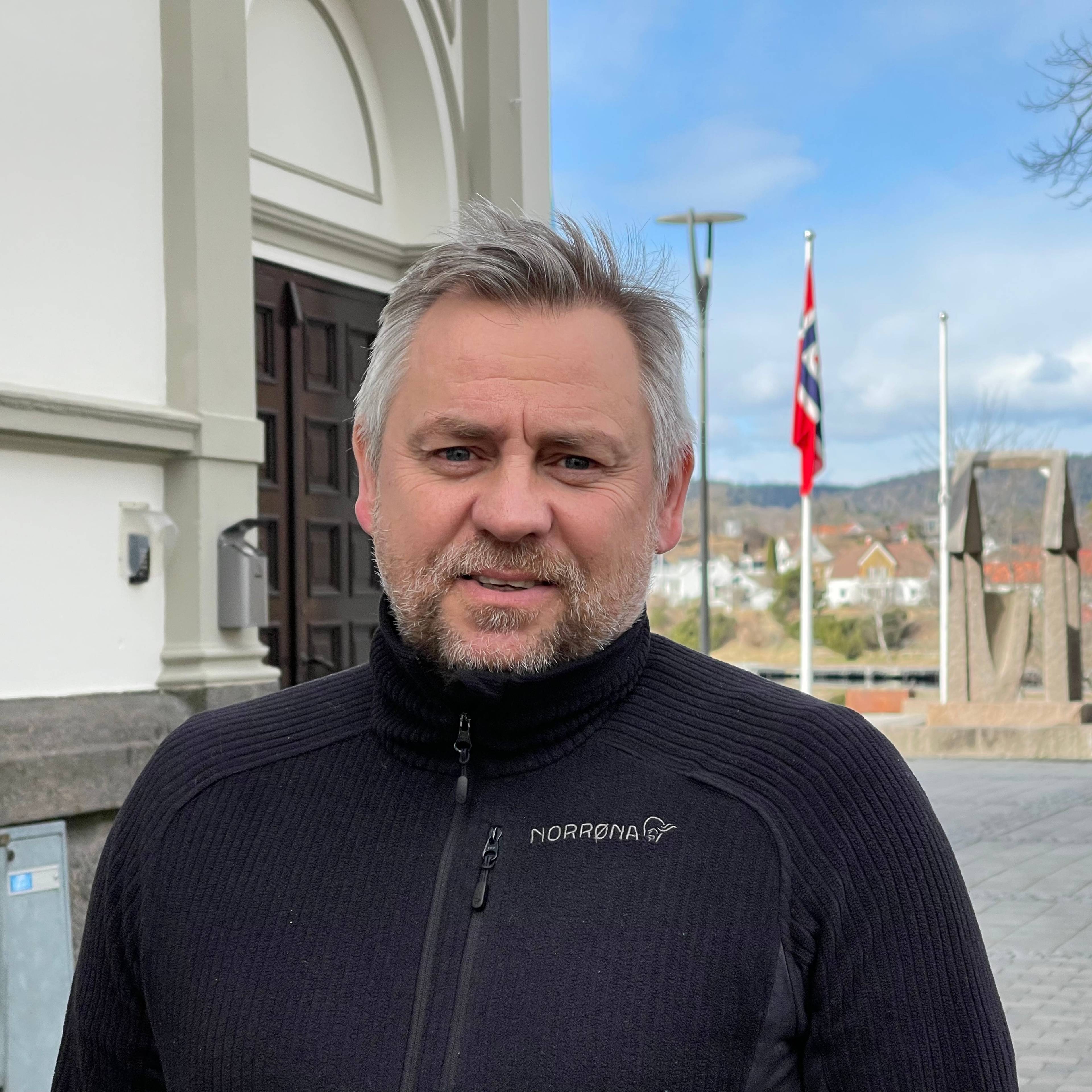En mann med grått hår står foran en bygning. I bakgrunnen er det et norsk flagg og et tettbebygd strøk. Mannen har på seg en svart genser. (Bildebeskrivelsen er laget av en KI-tjeneste)
