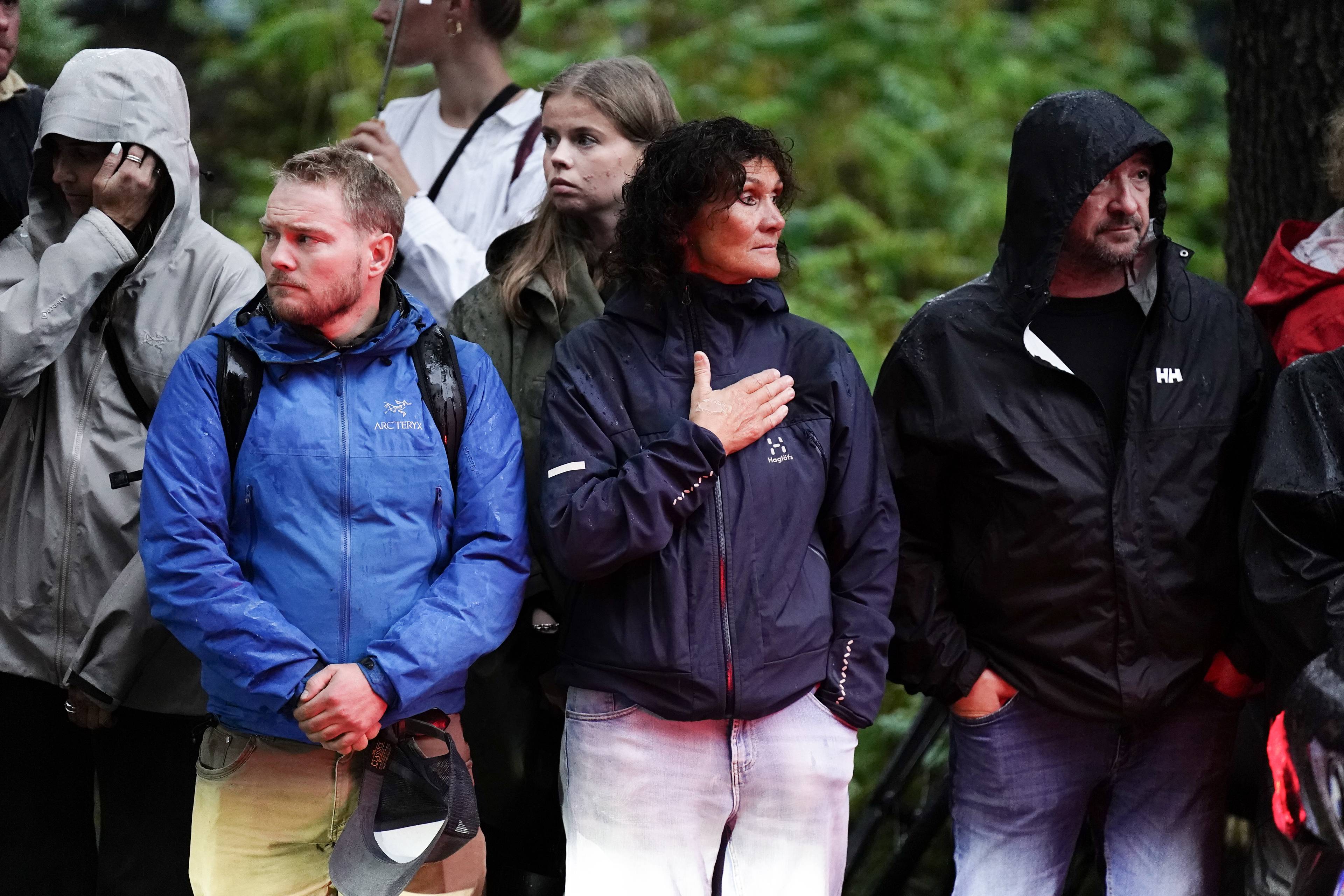 Personer står utendørs i regnvær. En kvinne i midten har hånden mot brystet. (Bildebeskrivelsen er laget av en KI-tjeneste)