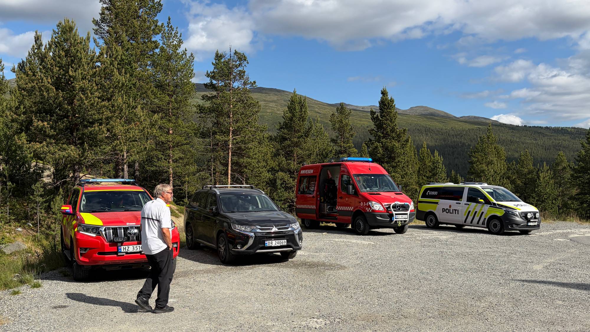 Nødetater på parkeringsplass i skogsområde. En person står ved siden av et rødt utrykningskjøretøy. 