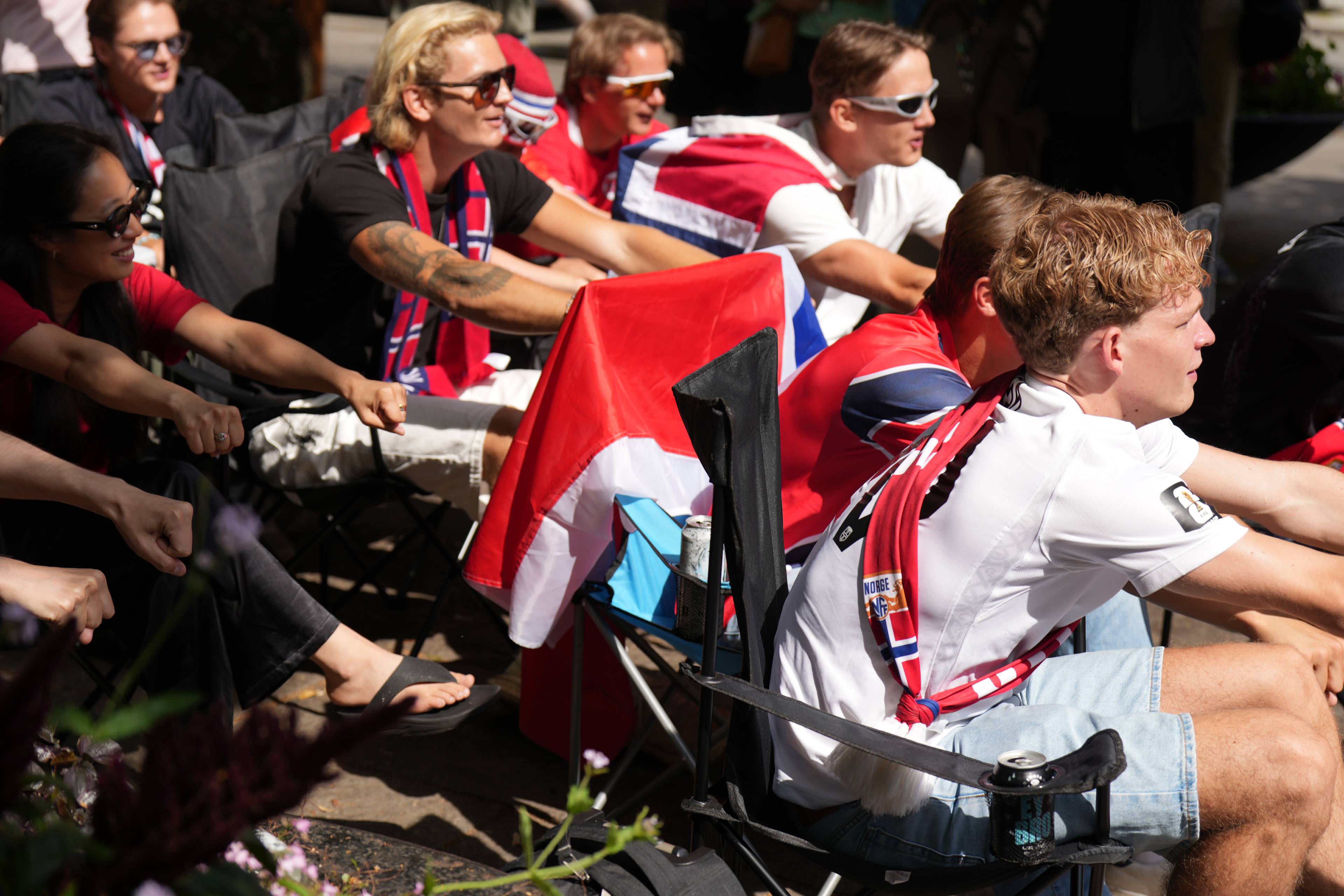 En gruppe mennesker sitter på campingstoler utendørs, kledd i norske farger og med norske flagg. (Bildebeskrivelsen er laget av en KI-tjeneste)