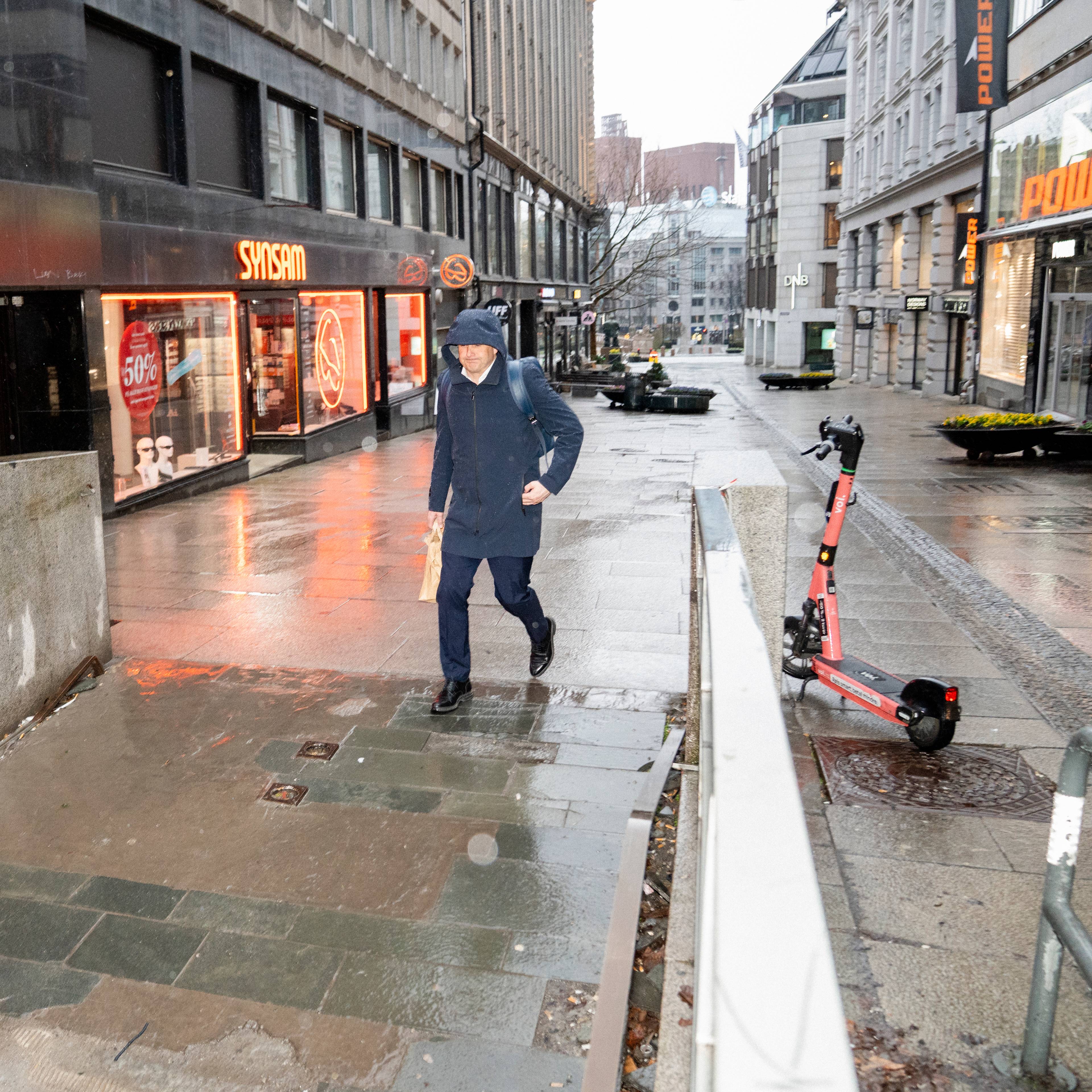 En mann i mørk frakk og bukse går over en våt gate. Det er neonreklame på en butikkvegg i bakgrunnen. En elektrisk sparkesykkel står på fortauet til høyre. (Bildebeskrivelsen er laget av en KI-tjeneste)