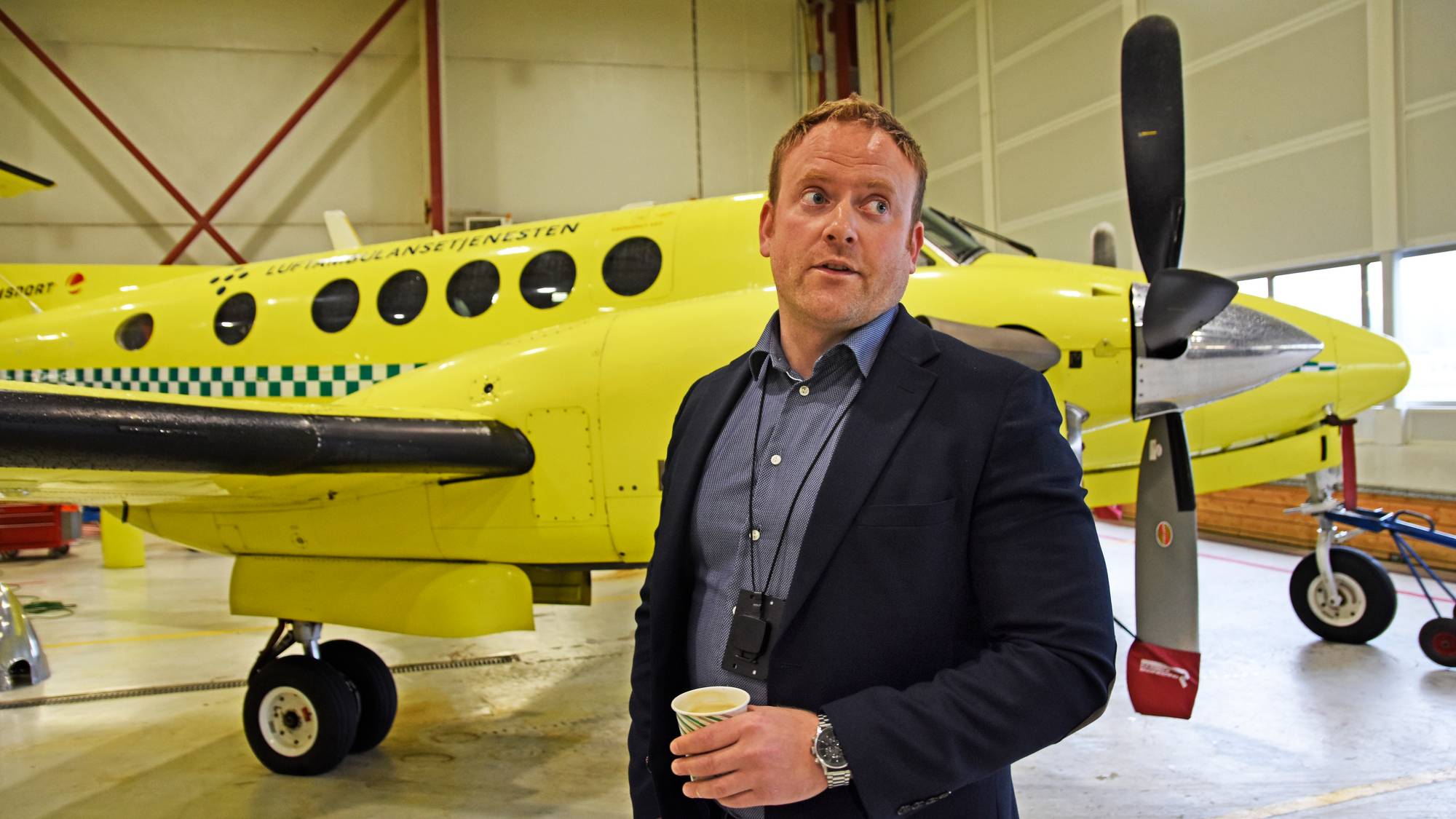 En mann i sort frakk innendørs i en hall. Et gult fly i bakgrunn. 
