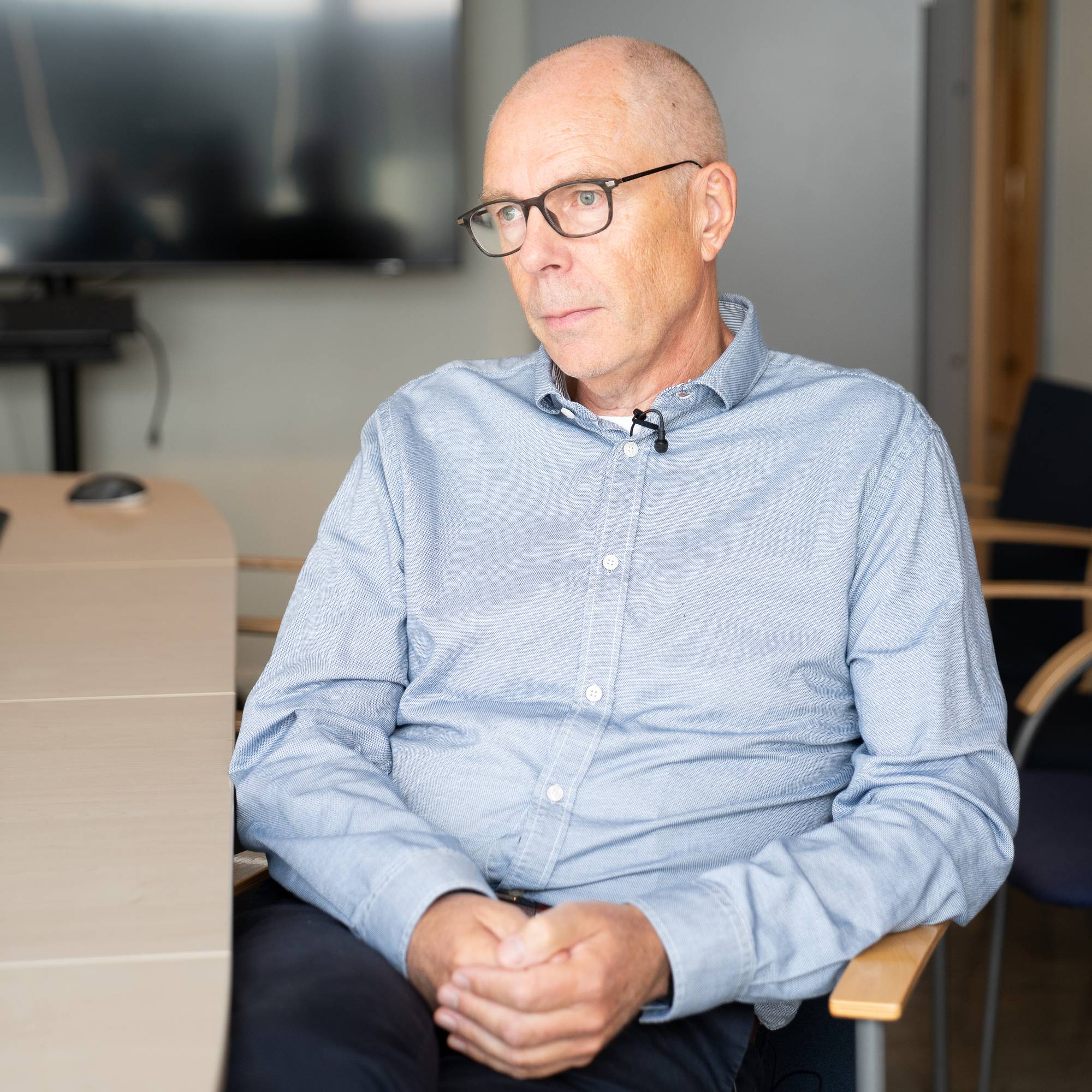 Mann med briller og lyseblå skjorte sitter ved et møtebord med en mikrofon festet til skjorta.