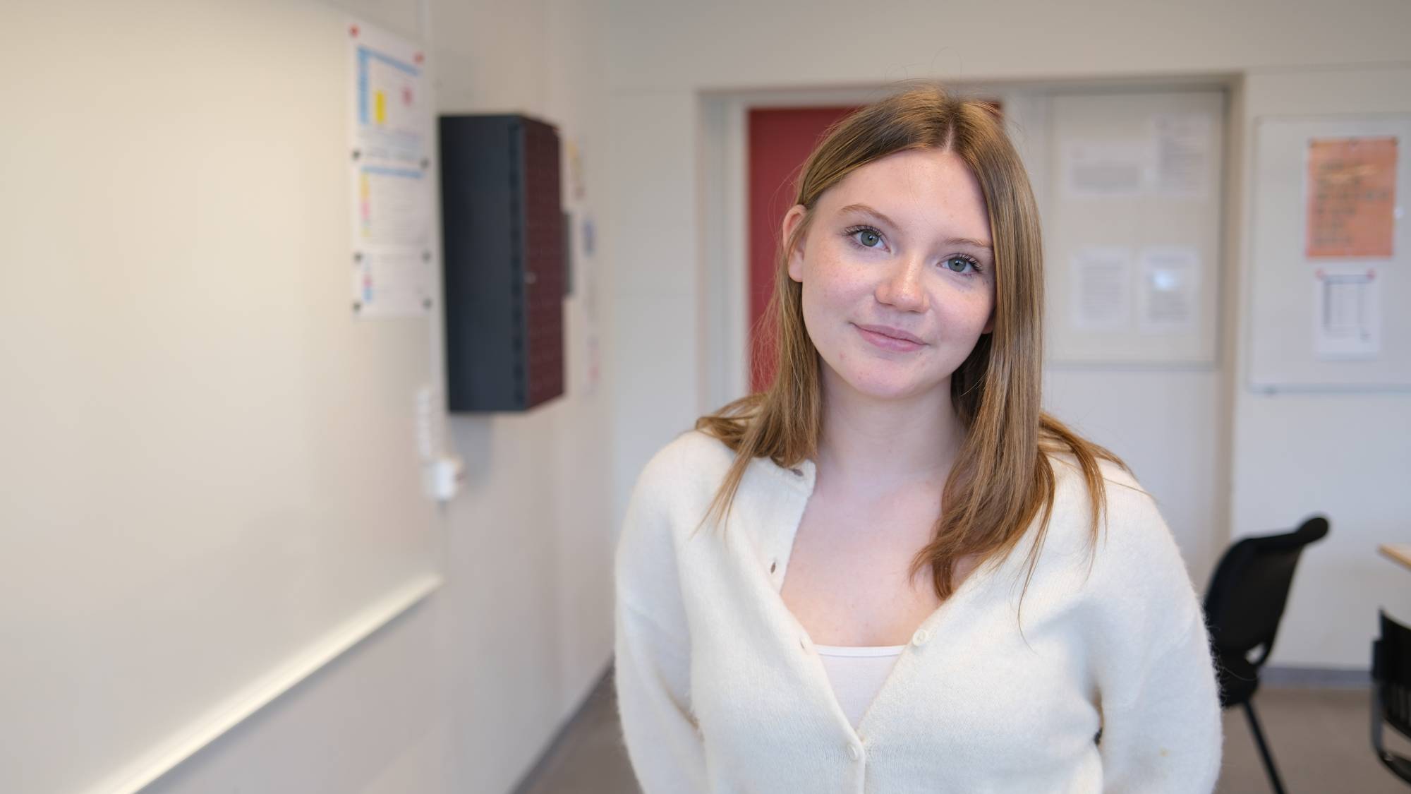 En ung kvinne med brunt hår i en hvit cardigan står i et klasserom og ser mot kameraet. (Bildebeskrivelsen er laget av en KI-tjeneste)