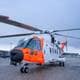 Heiko Junge / NTB En redningshelikopter står på bakken med oransje og hvite detaljer. Helikopteret har synlige roterblader og er parkert på en flyplass. I bakgrunnen sees en militærhelikopter. Omgivelsene er preget av snø og en gråhvit himmel.