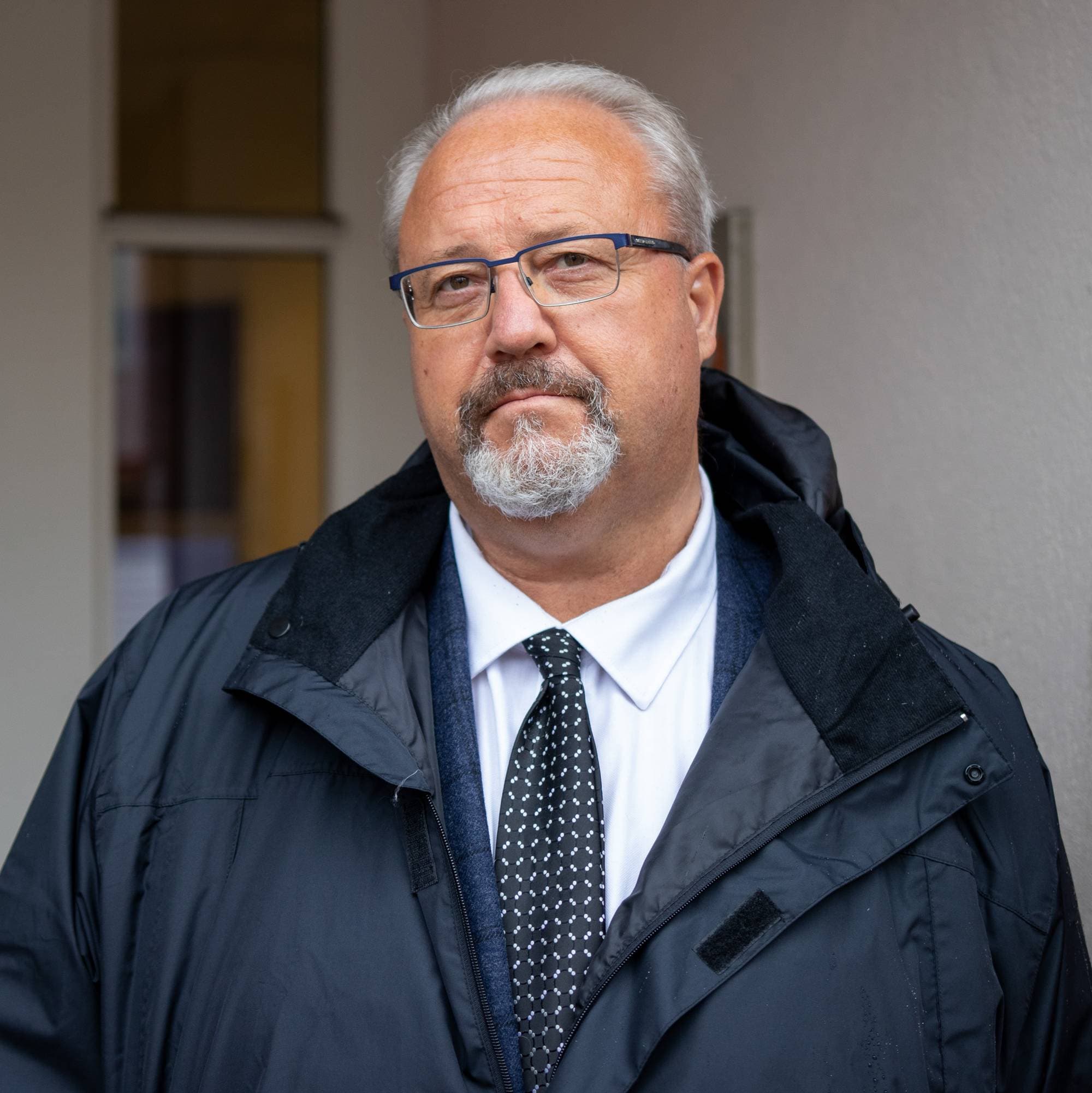 En mann med briller, skjegg og dress står utendørs foran et bygg