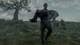 Foto: Steffan Hill / HBO A man carries a heavy man down a green hill. Several horses in the background.